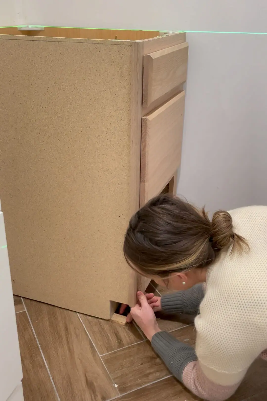 A woman adjusting wooden shims under the corner of an unfinished bathroom cabinet to ensure it is level. The green laser line highlights alignment across the wall and cabinet.