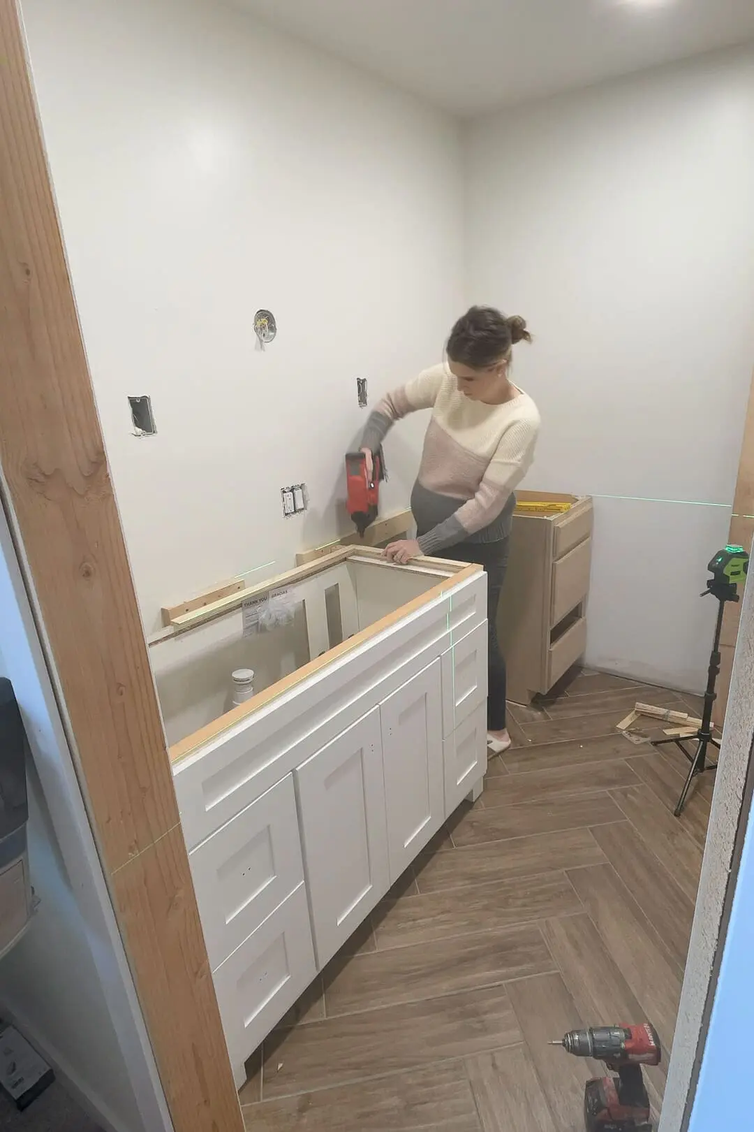 Woman in a striped sweater using a nail gun to install a support board for a white shaker bathroom cabinet. A green laser level projects guidelines onto the wall, and tools are scattered on the herringbone floor.