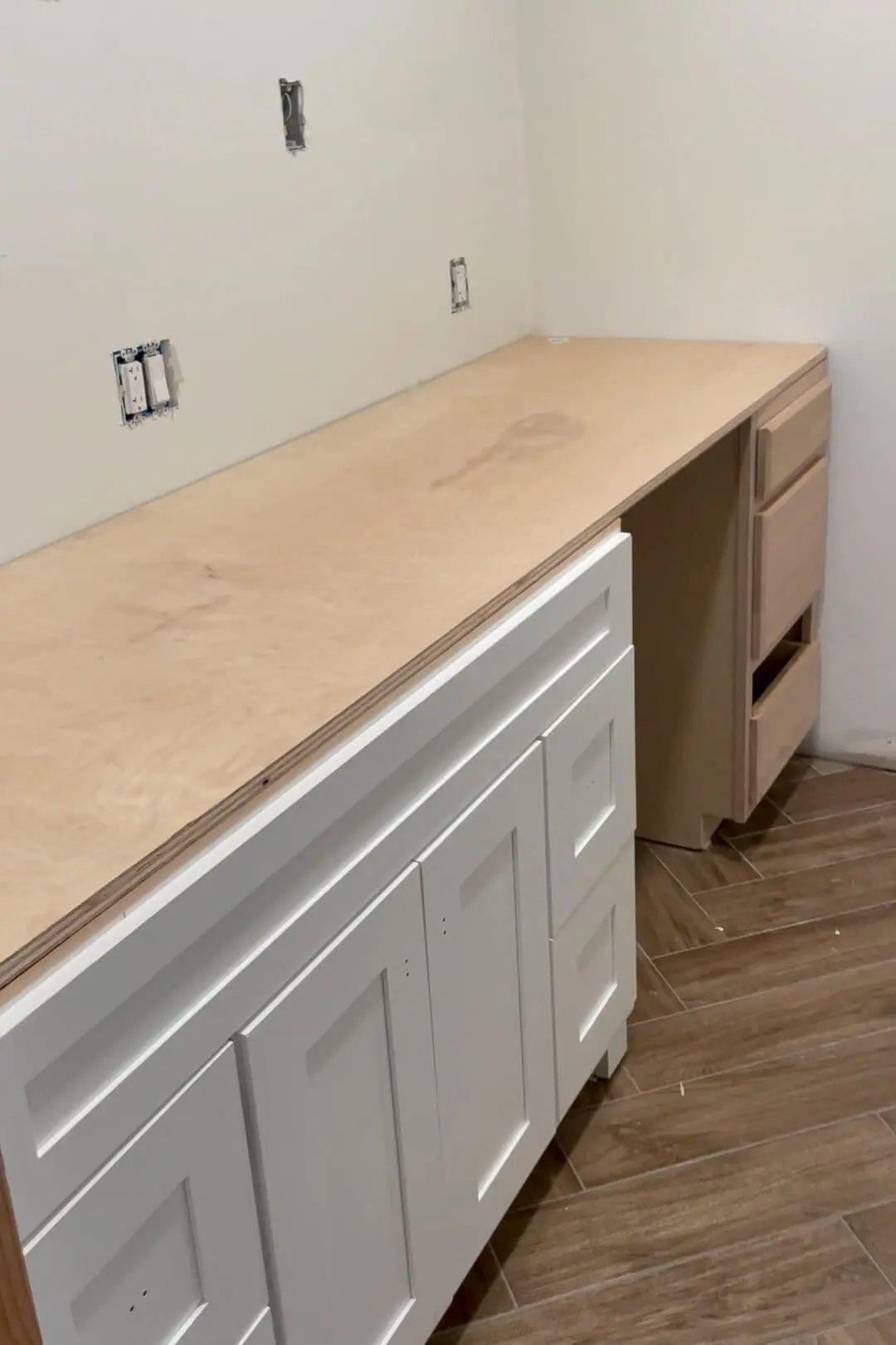 Partially installed plywood countertop over white shaker-style bathroom cabinets and an unfinished wooden cabinet. The clean, minimal space features wood-look herringbone flooring and prepped electrical outlets.