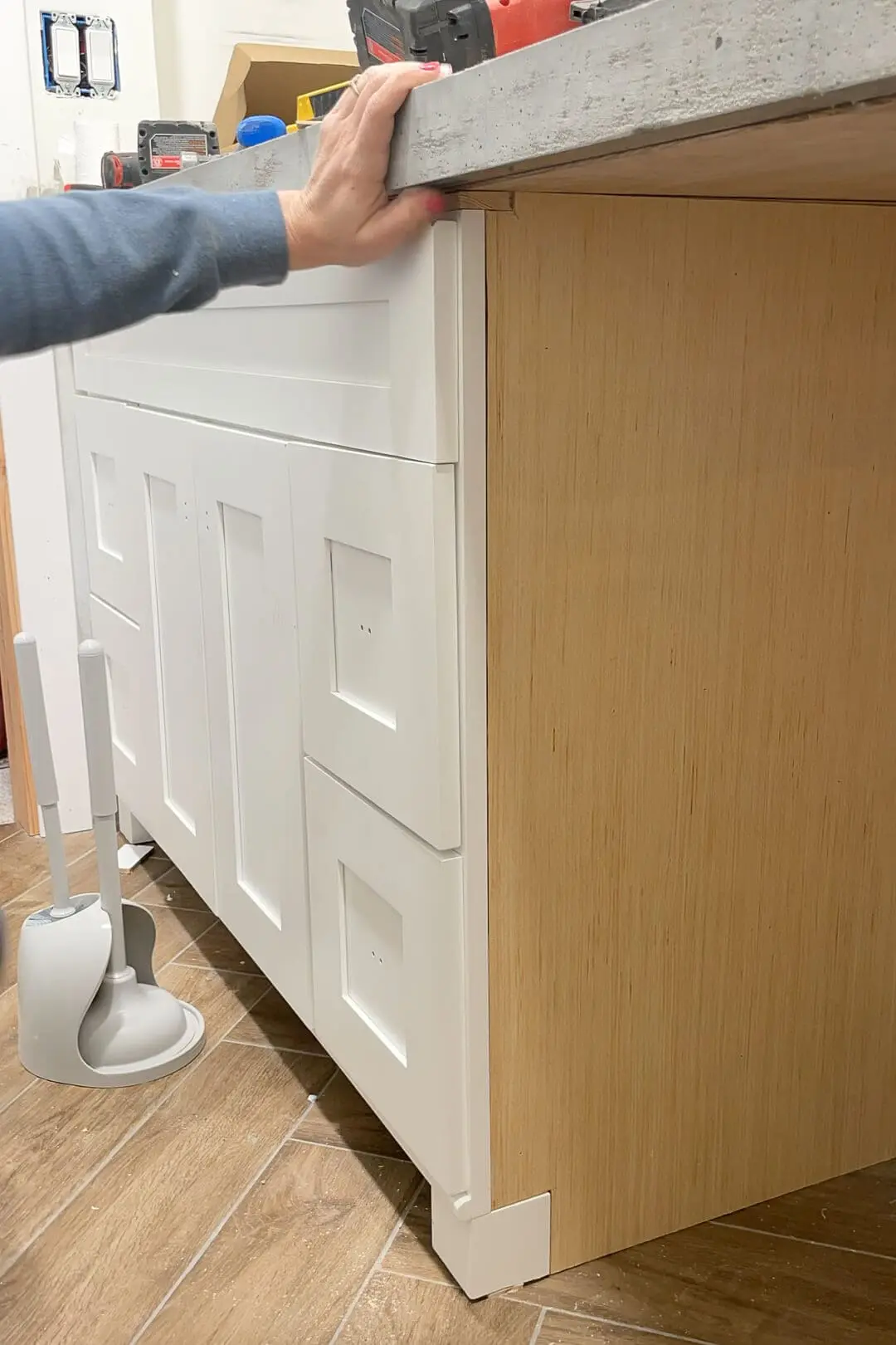 Close-up view of a hand holding onto a concrete countertop edge above a white shaker cabinet with an installed wood veneer panel on the side, during a bathroom renovation.