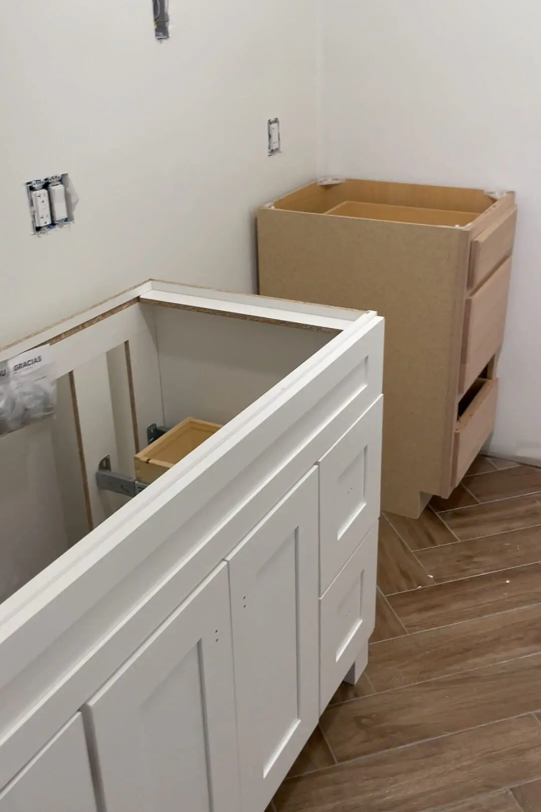 In-progress bathroom renovation featuring partially installed white shaker-style vanity cabinet and unfinished wood kitchen drawer cabinet. Electrical outlets and wall cutouts are visible on a clean white wall, with wood-look floor tiles laid in a herringbone pattern.