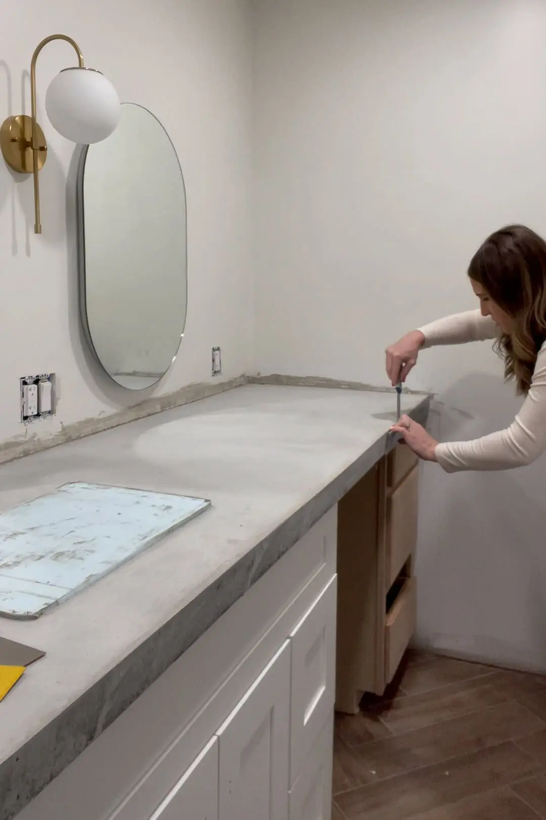 Woman smoothing and leveling a freshly installed concrete countertop on a white shaker-style custom bathroom vanity during a bathroom renovation. An oval mirror, gold wall sconce, and unfinished wooden cabinet drawers are visible in the modern space.