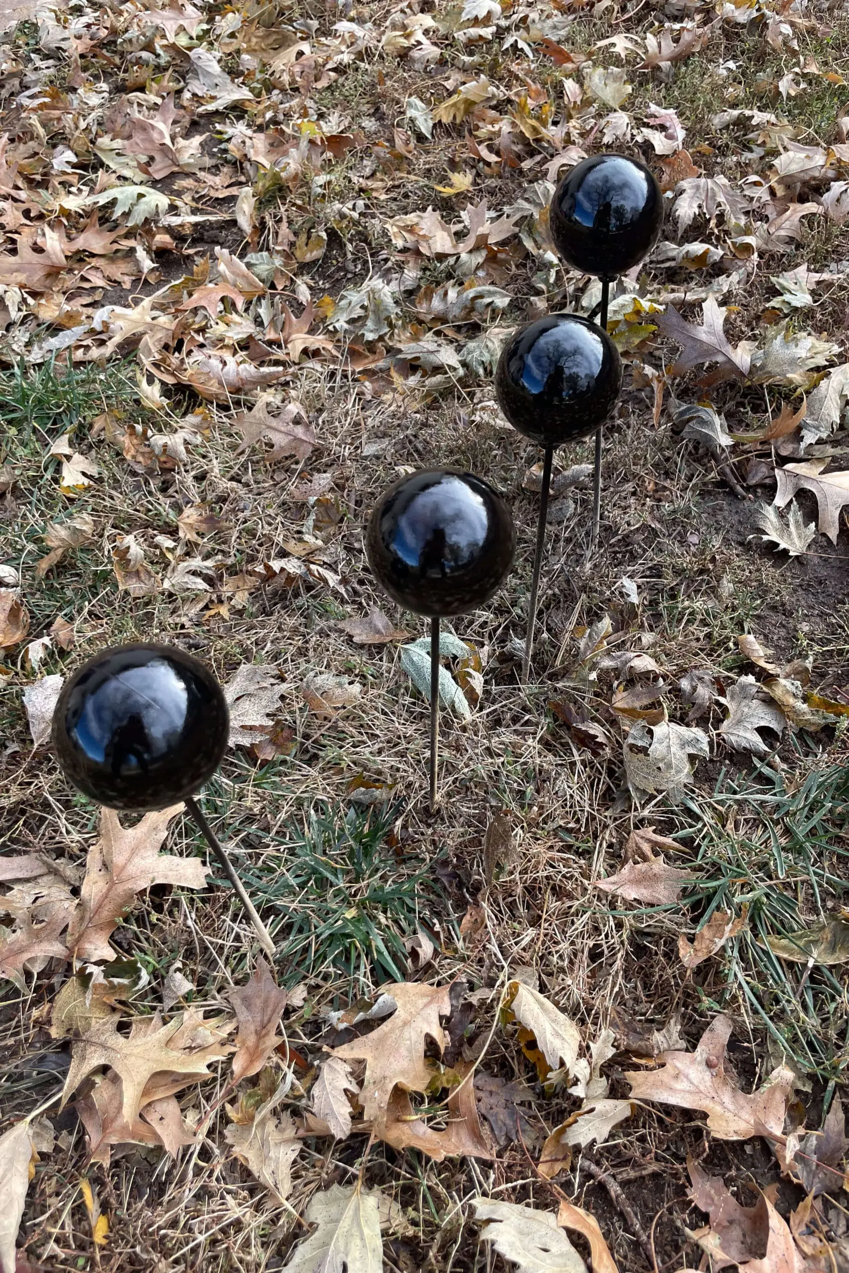 Four glossy black-painted glass ornaments on skewers displayed outdoors on a patch of grass surrounded by fallen leaves.