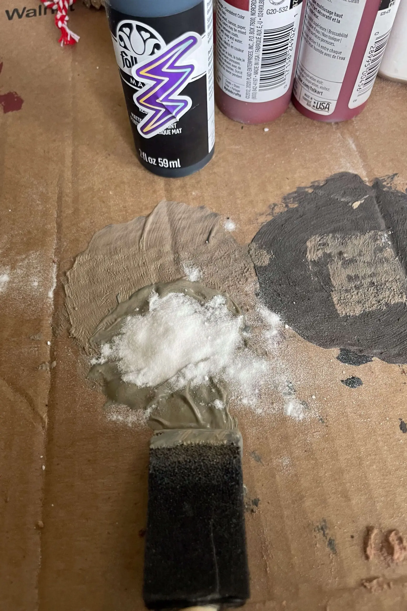 A close-up of gray paint mixed with baking soda on a cardboard surface, with a foam brush and FolkArt matte acrylic paint bottle nearby, showcasing a textured craft mixture.