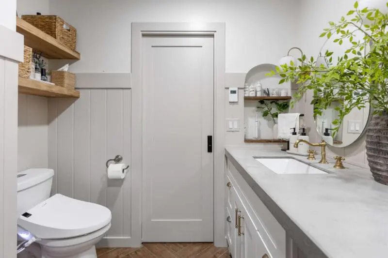 Modern bathroom featuring a white shaker vanity with a concrete countertop, gold faucet hardware, and an oval mirror. A built-in shelf with wooden open storage holds decorative baskets and toiletries. A smart toilet, light gray door, and paneling complete the clean, contemporary design.