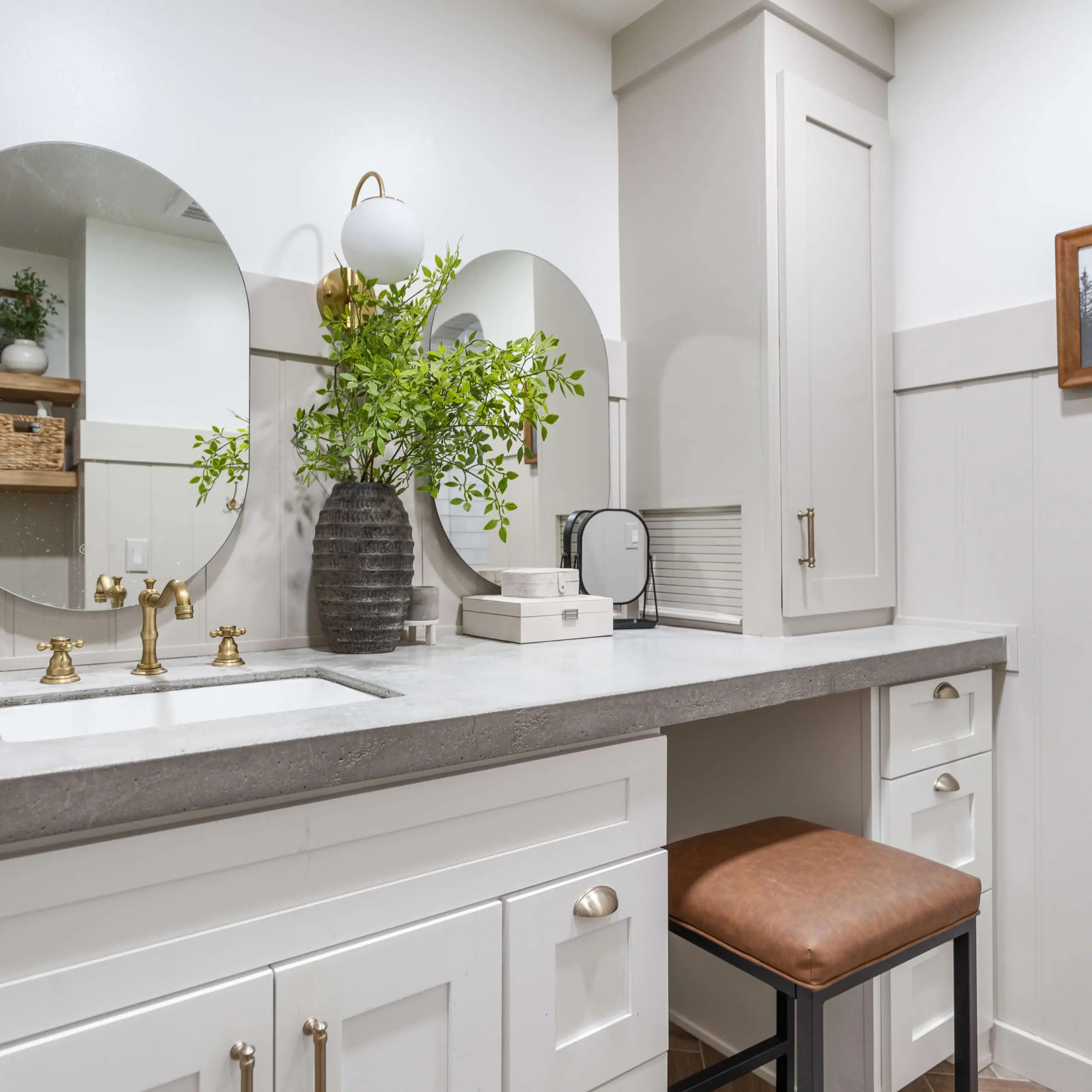 Modern bathroom vanity with white shaker cabinets, a concrete countertop, and gold hardware. Two round mirrors are mounted above the sink, flanked by a black vase with green branches. A sleek built-in cabinet provides additional storage, while a brown leather stool sits tucked neatly under the counter, completing the organized and stylish space.