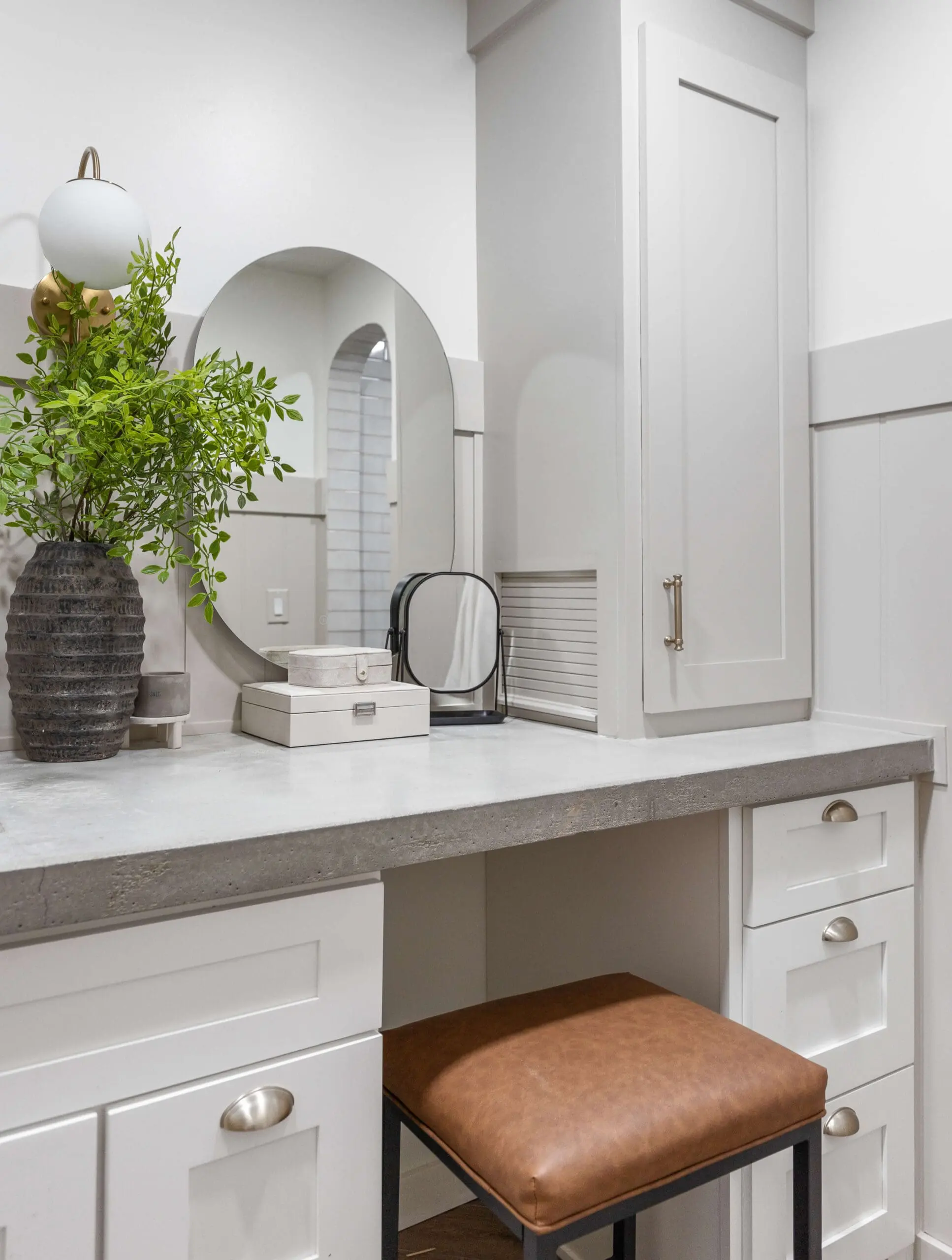 Modern bathroom vanity with white shaker cabinets, a concrete countertop, and gold hardware. Two round mirrors are mounted above the sink, flanked by a black vase with green branches. A sleek built-in cabinet provides additional storage, while a brown leather stool sits tucked neatly under the counter, completing the organized and stylish space.