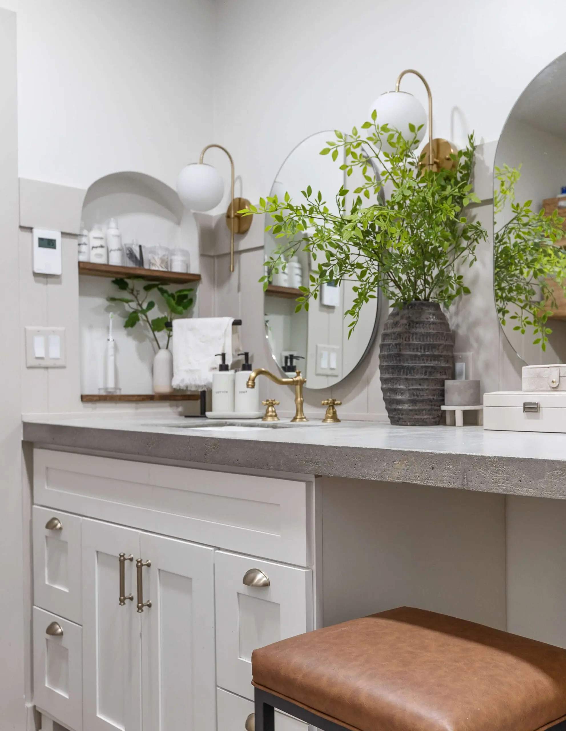Close-up view of a modern bathroom vanity featuring white shaker cabinets, a thick concrete countertop, and gold faucet hardware. A black vase with lush green branches adds a natural touch, while a round mirror and gold wall-mounted sconces enhance the clean, stylish design. A brown leather stool sits tucked under the countertop, and open shelving holds organized toiletries and decor.