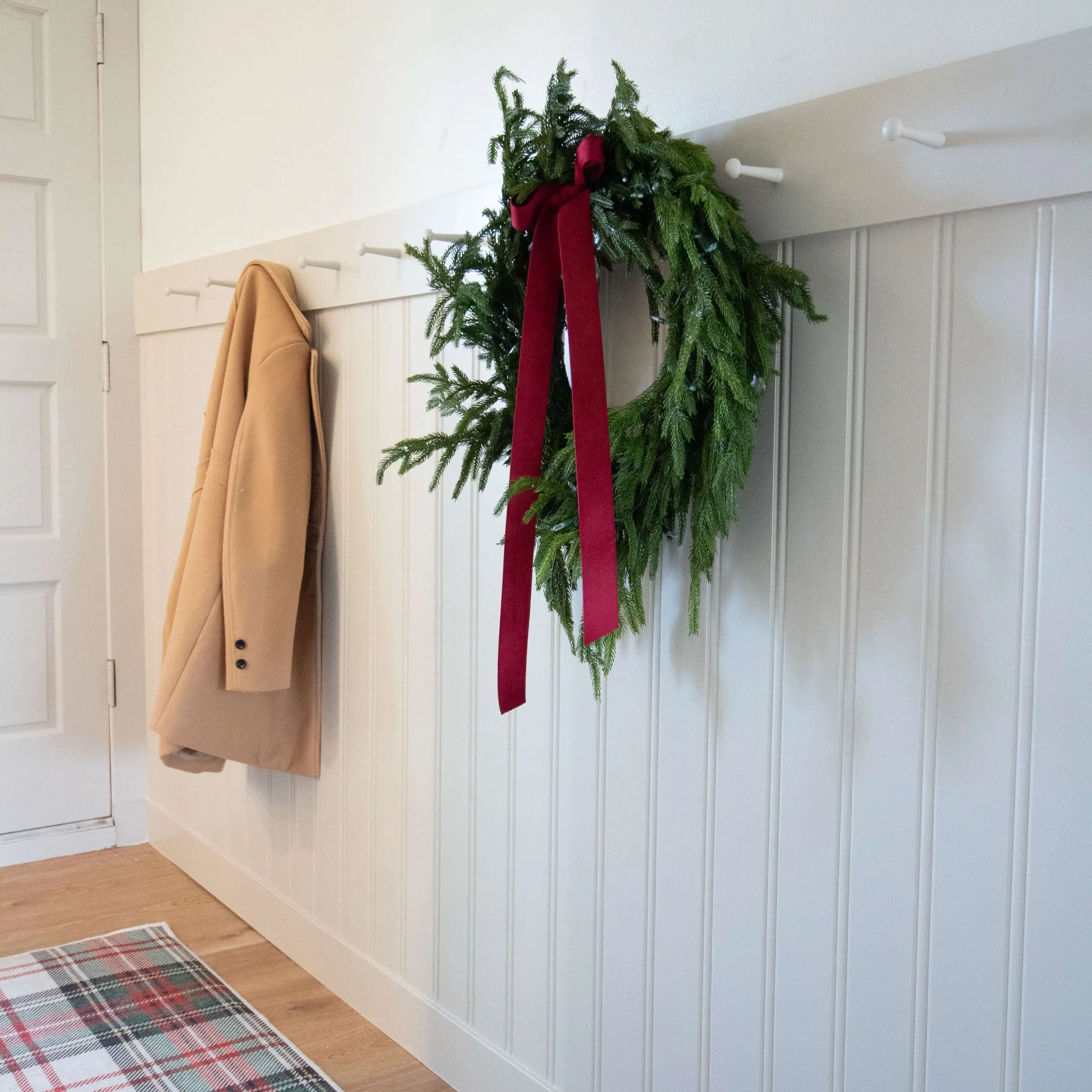 Close-up of a white wall coat rack with a sleek, minimalist design, showcasing a beige coat and a decorative holiday wreath with greenery and a red ribbon.