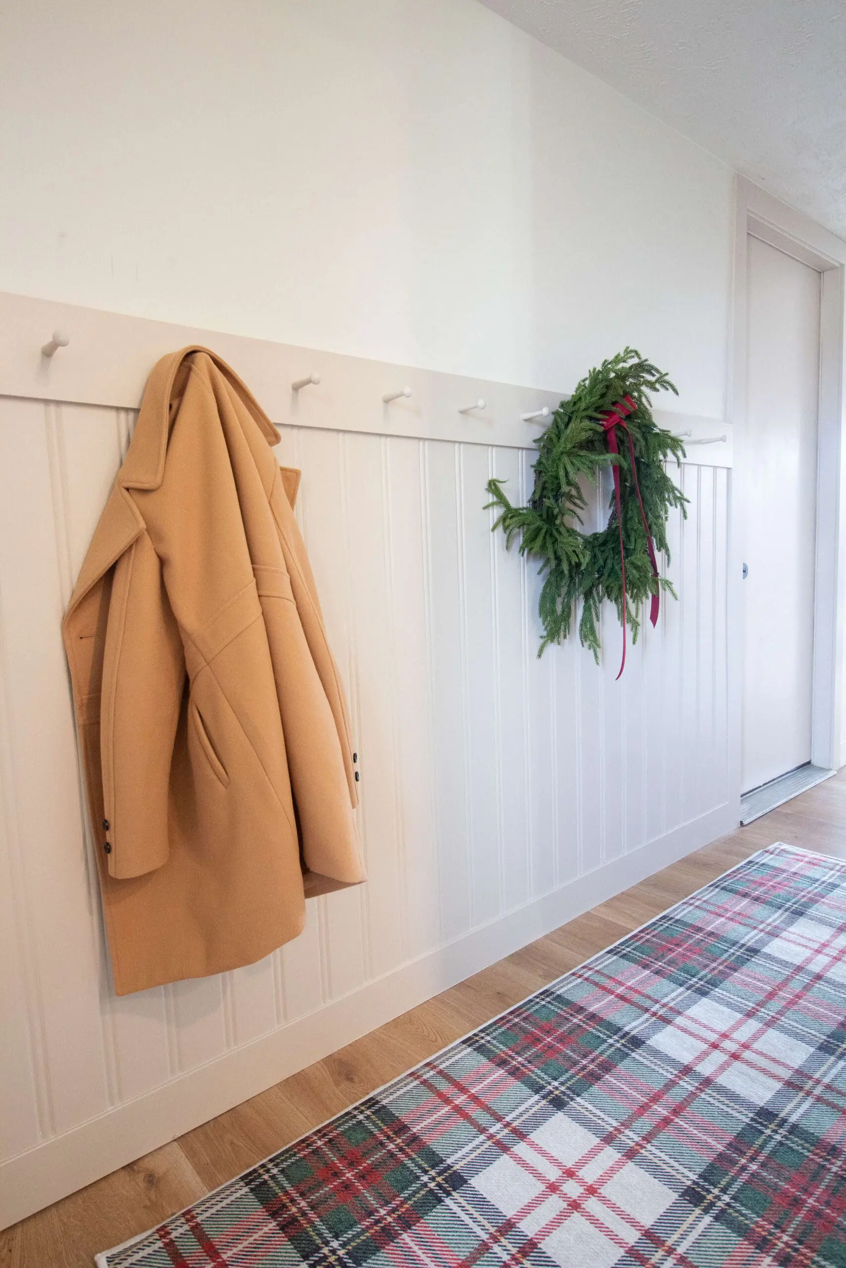 A bright hallway featuring a white wall coat rack with a beige coat and a green holiday wreath, set against beadboard paneling and a plaid area rug.