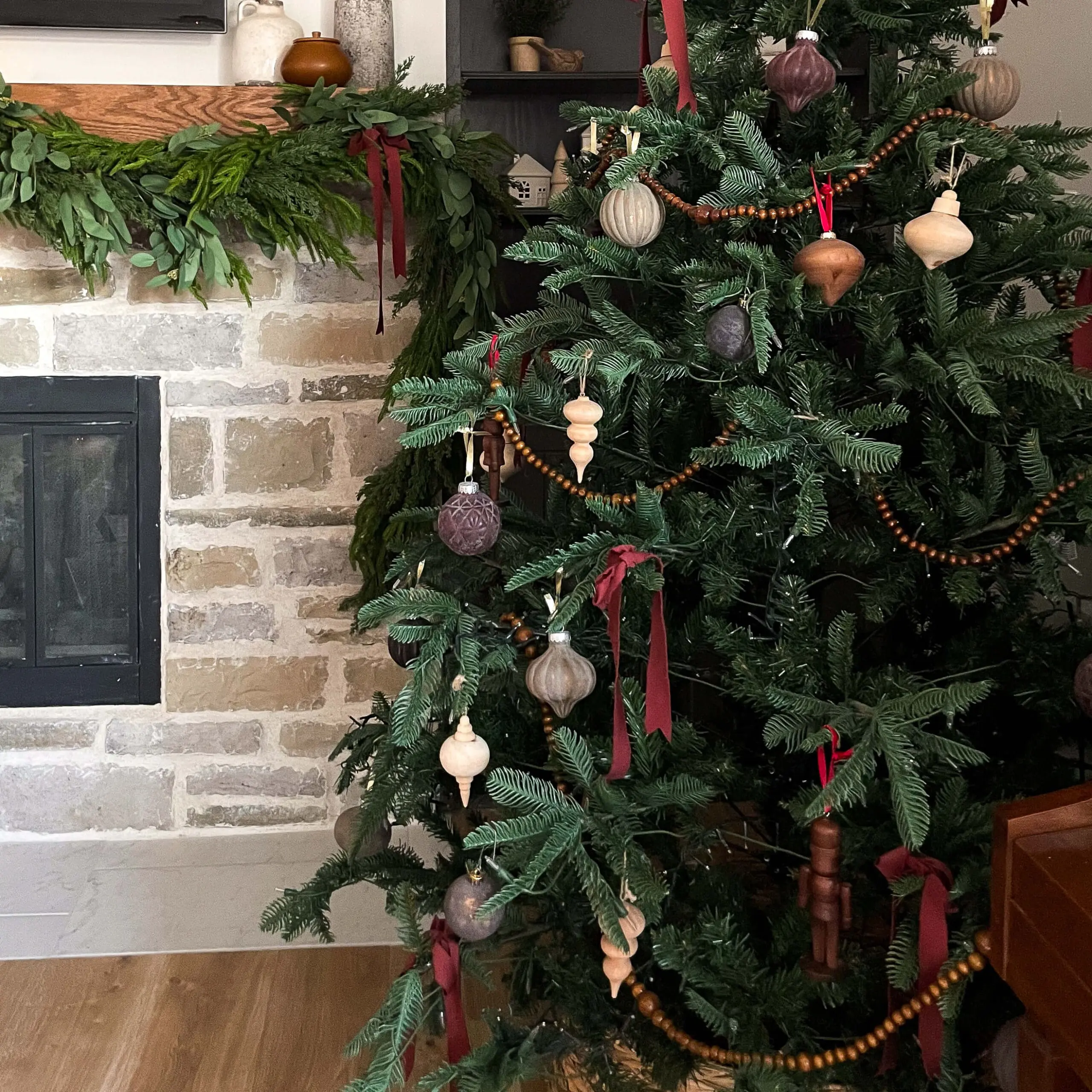 A cozy living room decorated for Christmas with a natural-themed artificial tree featuring DIY flocked ornaments, wooden ornaments, deep red ribbons, and wooden bead garland. The tree stands next to a rustic stone fireplace adorned with a lush green garland and minimalistic decor on the mantel.
