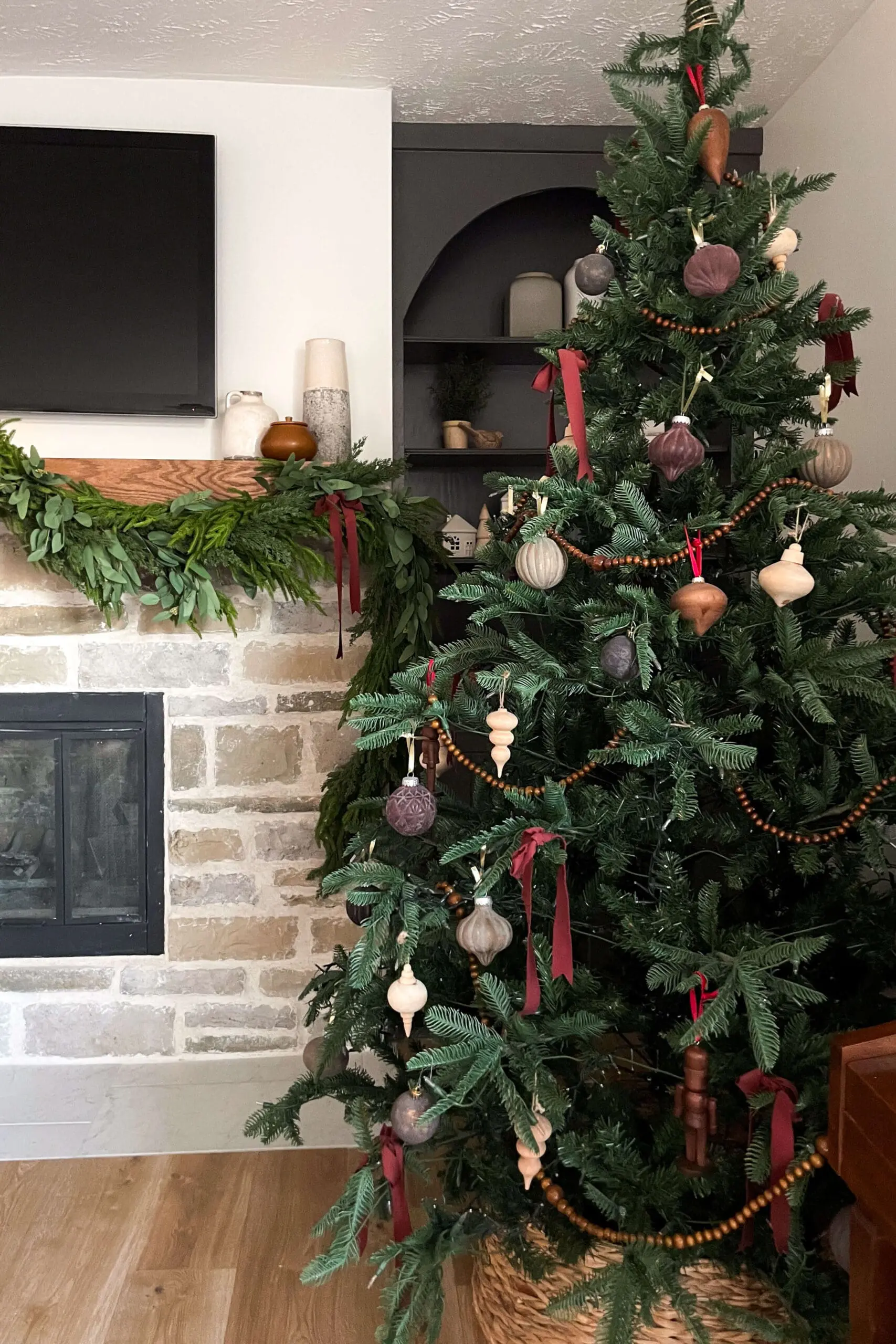 A warm Christmas living room setup showcasing an artificial tree adorned with wooden ornaments, muted red ribbons, and a wooden bead garland. The tree is paired with a rustic stone fireplace dressed in a greenery garland, complementing the natural aesthetic of the room.