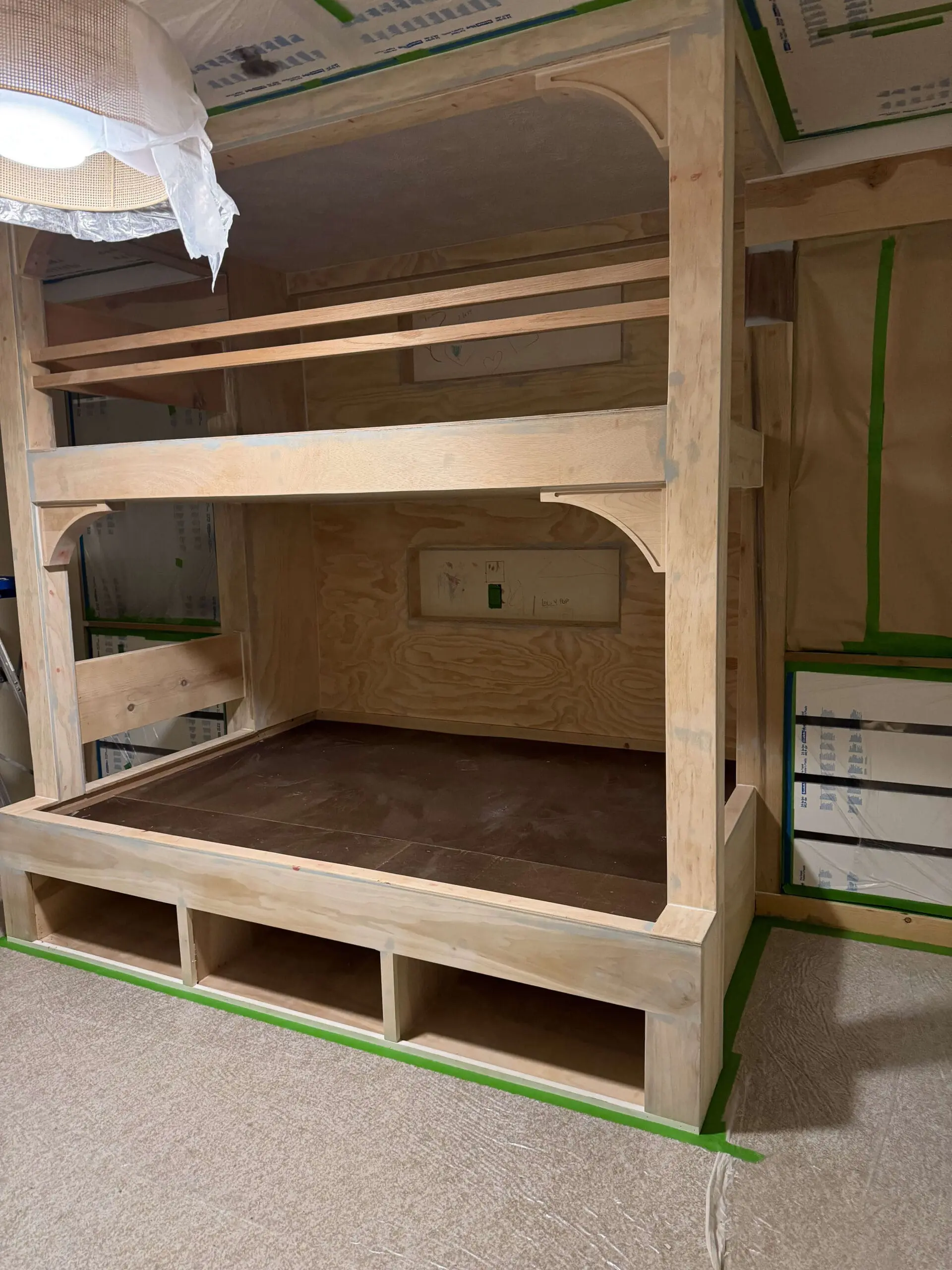 A custom-built wooden bunk bed frame in progress, featuring built-in cubby storage at the base and an arched design for the support beams, with all surfaces prepped and taped for painting.

