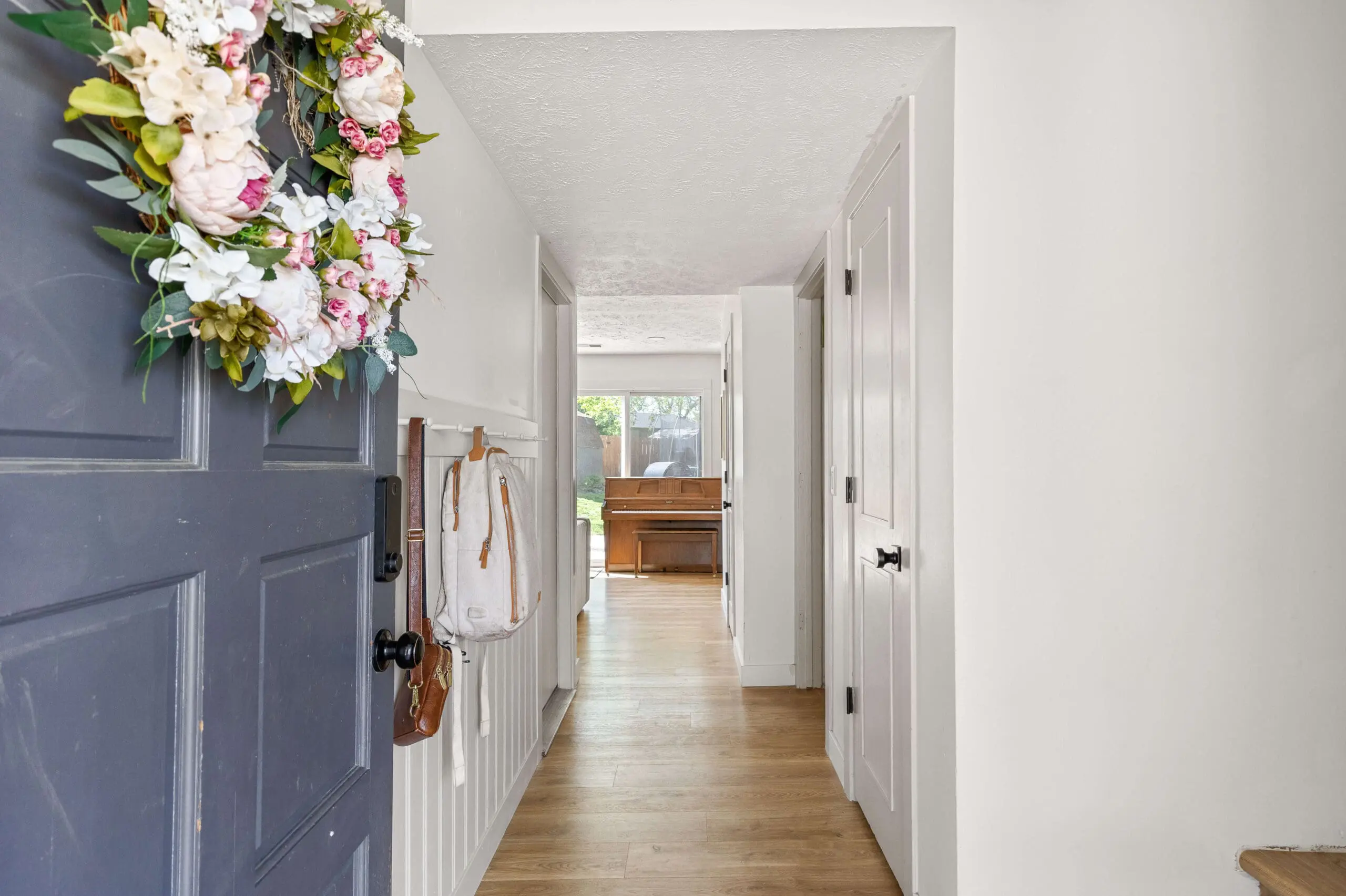 View into a modern farmhouse-inspired entry featuring a navy front door with a floral wreath, vertical board and batten wall trim, and backpack storage on wall hooks.