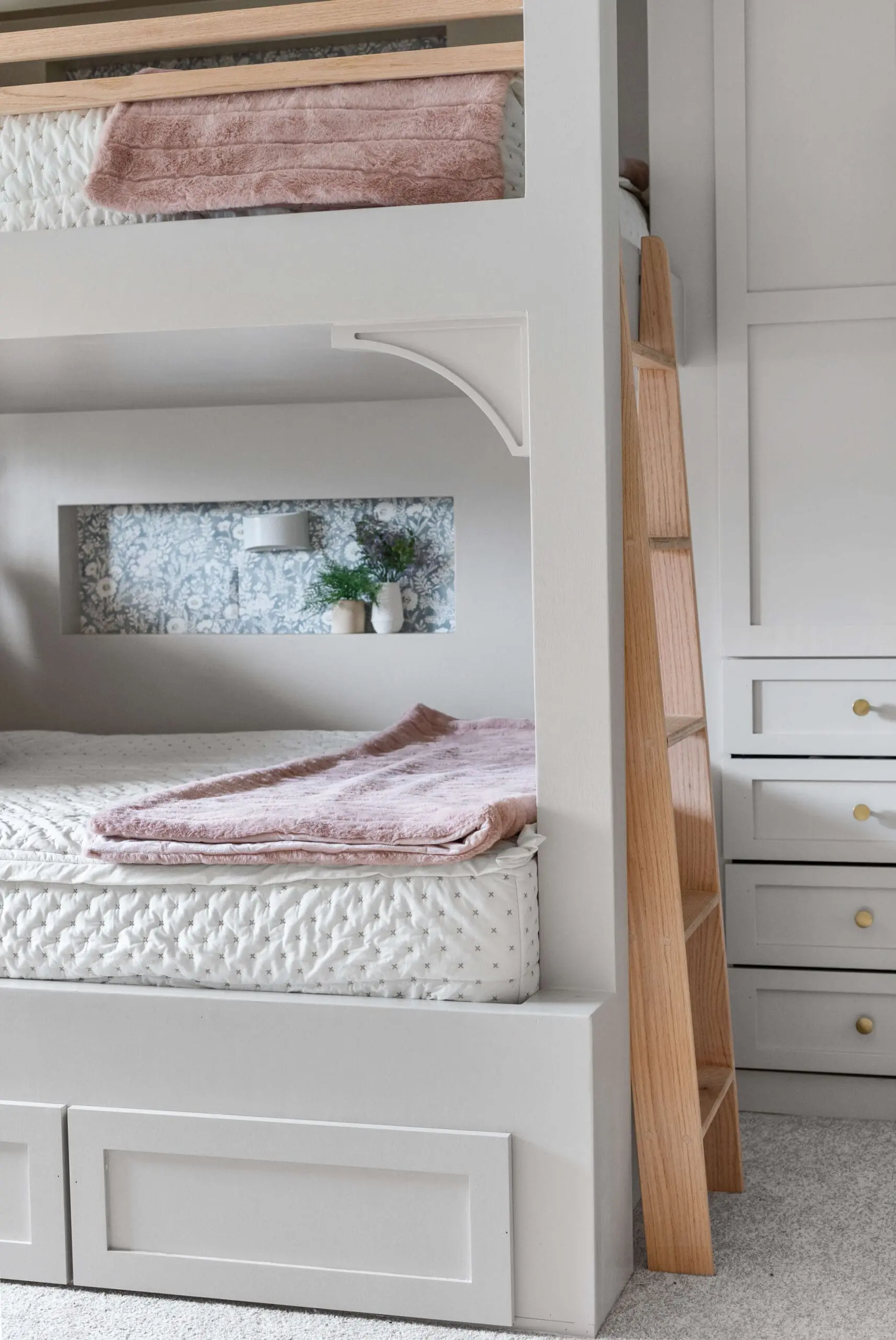 Charming bunk bed setup with light gray built-ins, a wooden ladder, and cozy bedding featuring soft pink blankets. The lower bunk includes a built-in shelf with floral wallpaper, small decor accents, and a sconce for added functionality and style.