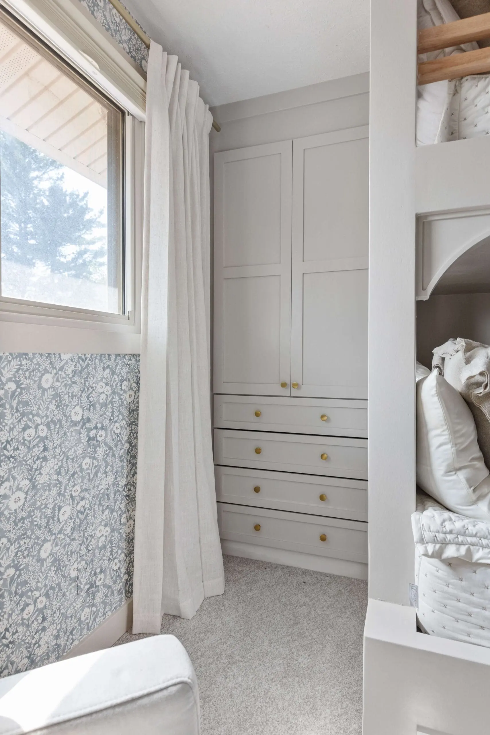 A cozy corner featuring built-in white shaker-style cabinets with matte gold knobs, framed by a window dressed in soft white curtains and blue floral wallpaper. A partial view of a custom bunk bed with plush bedding adds warmth and functionality to the space.