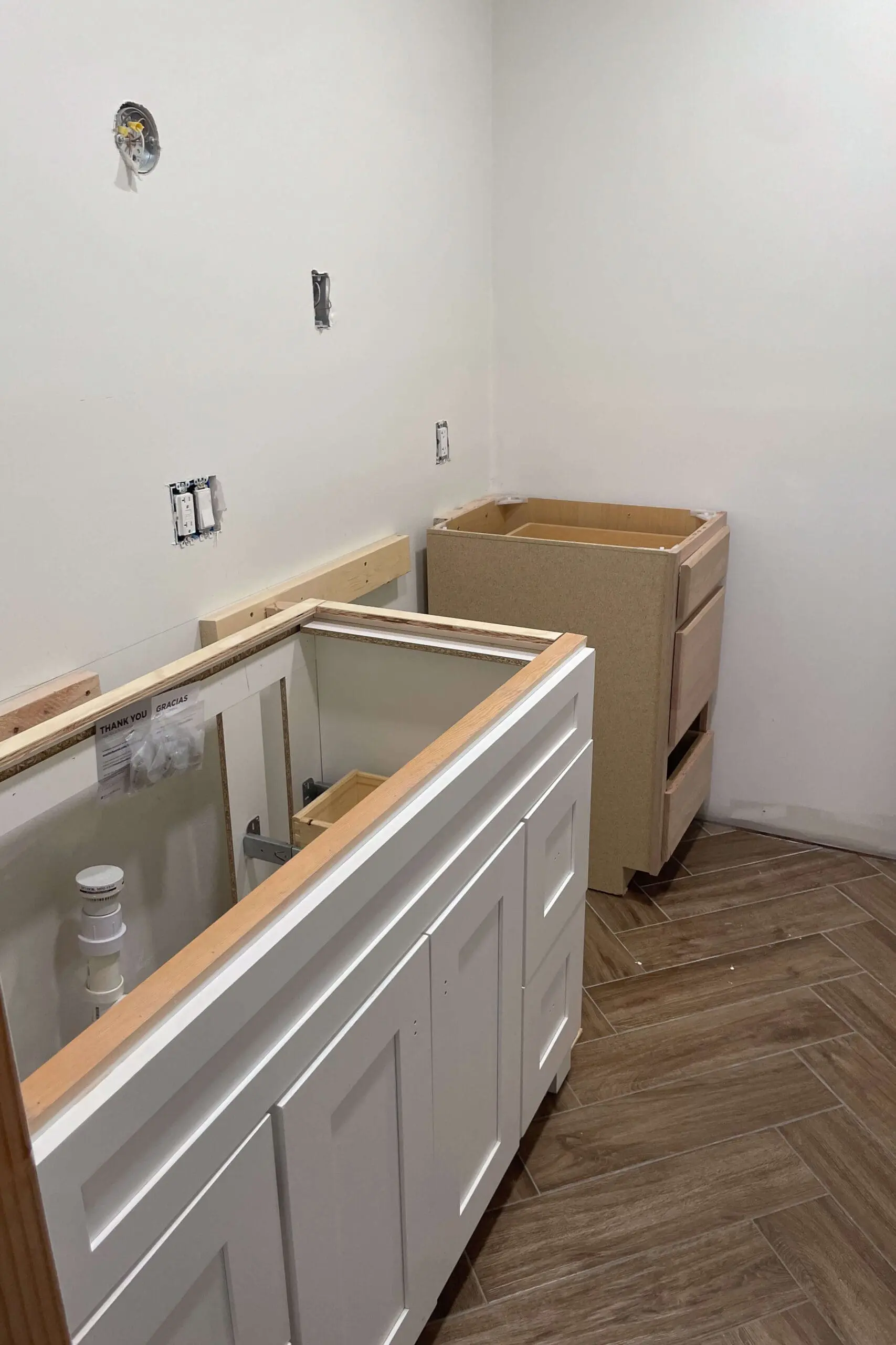 Bathroom renovation progress showing a white shaker-style cabinet and an unfinished wooden cabinet. Support boards are mounted to the wall, and visible plumbing is ready for the sink installation.