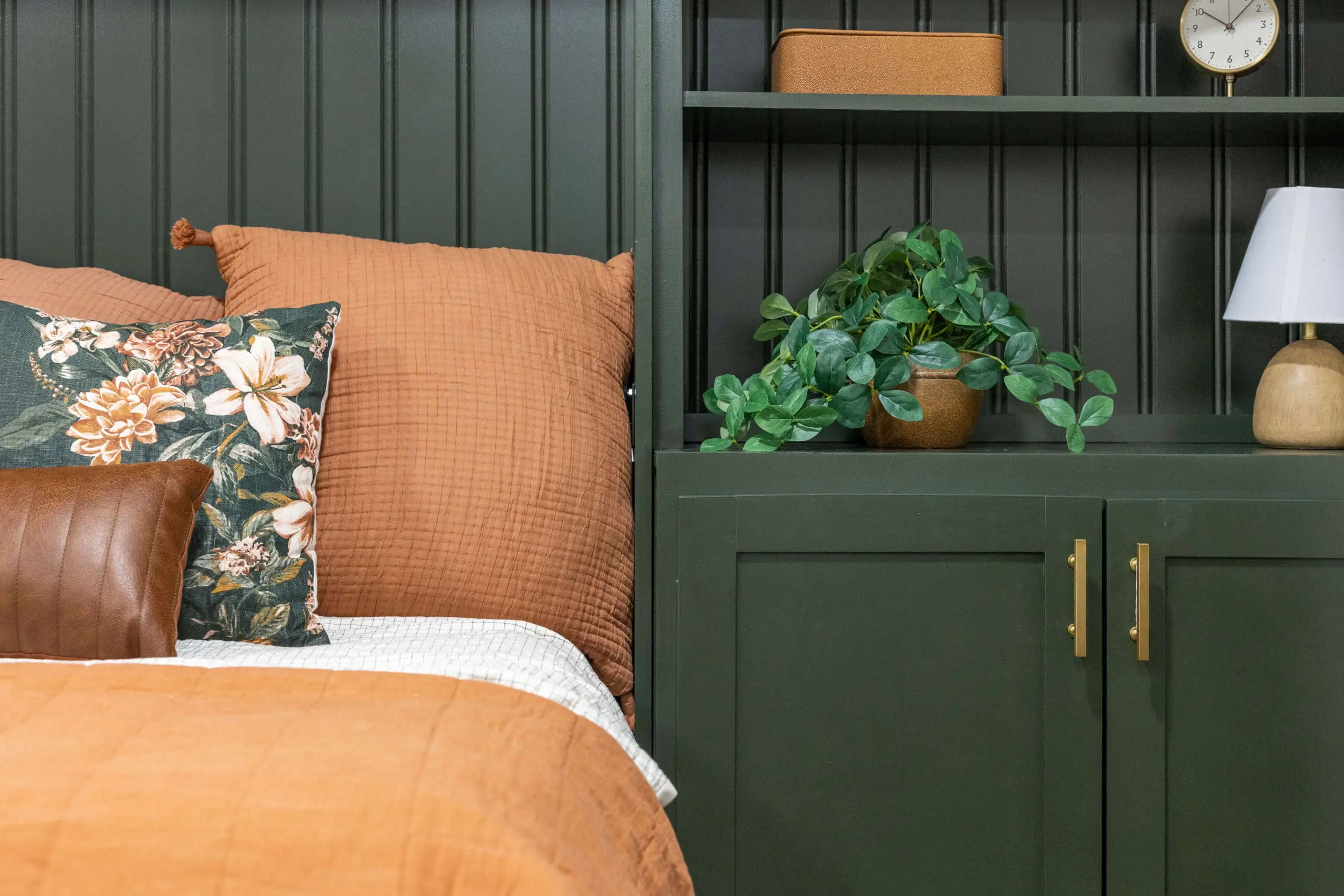 Cozy bedroom corner with deep green built-in cabinetry and beadboard detail, complemented by warm-toned bedding in orange and floral patterns. A leather accent pillow and greenery in a terracotta pot add texture, with small decorative accents like a lamp and clock on the shelving.