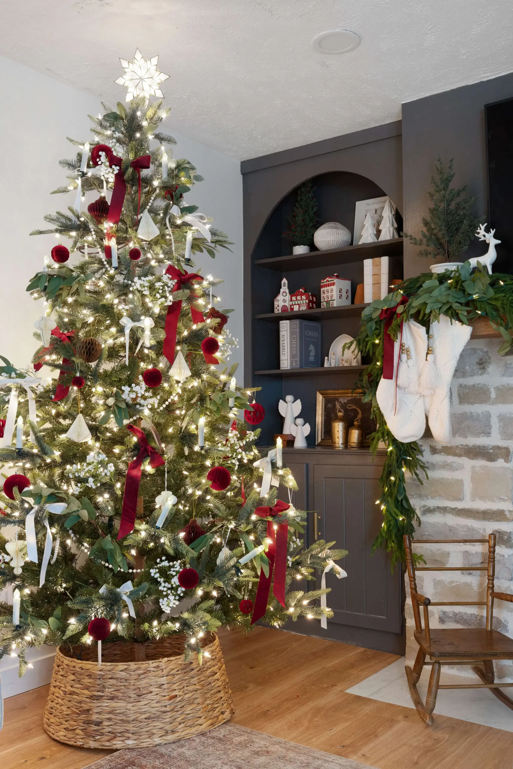 A festive Christmas tree decorated with classic red and white accents, including velvet bows, hanging bells, and faux white berries. The tree glows with warm lights and features a glowing star tree topper. The background includes a stone fireplace draped with garland and stockings, alongside a dark gray built-in shelf decorated with miniature houses, ceramic trees, and holiday greenery.