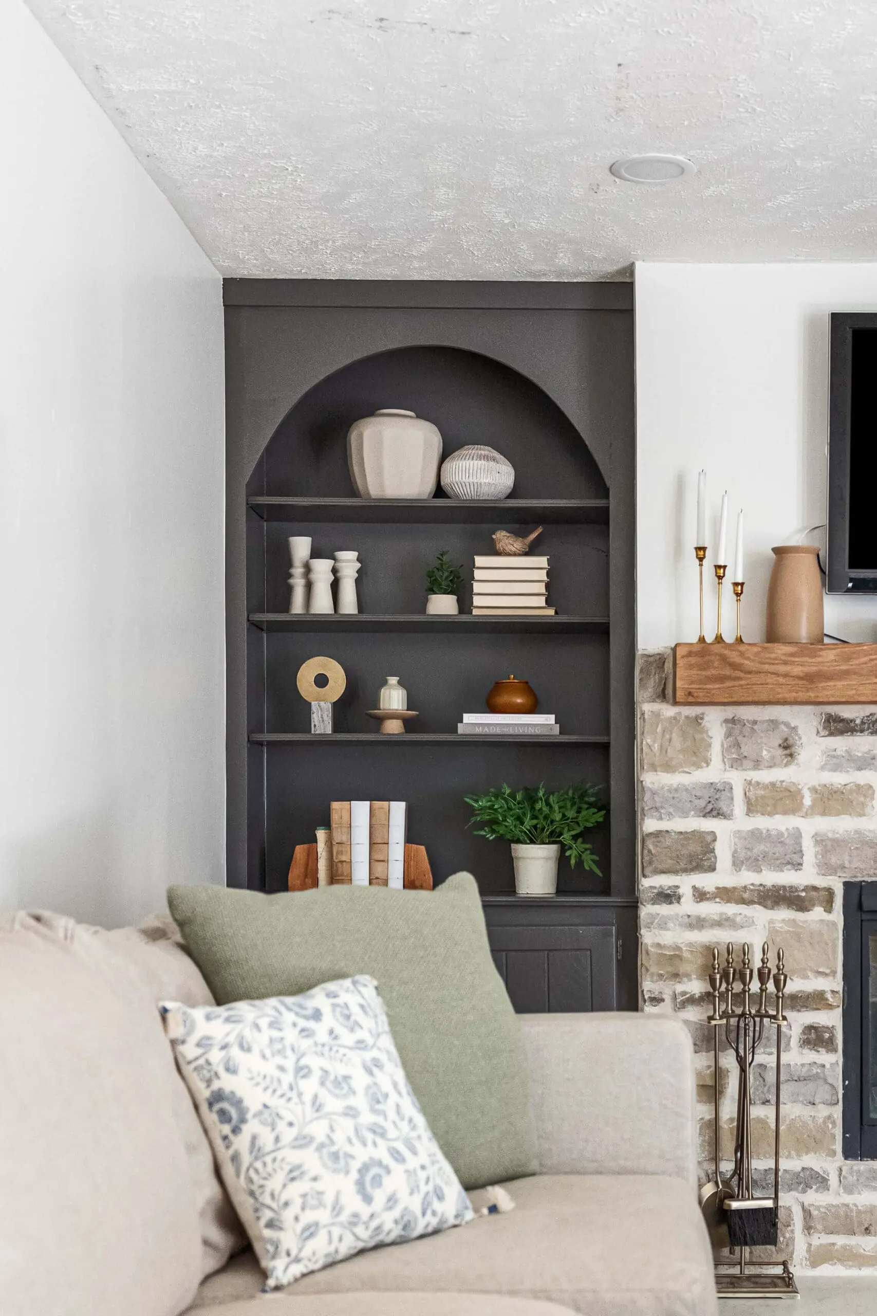 A finished built-in arched shelving unit painted in a deep gray, styled with neutral-toned decor, books, and greenery. Positioned next to a stone fireplace with a wooden mantel, this cozy living room corner includes a sofa with green and blue floral pillows for a polished and inviting look.