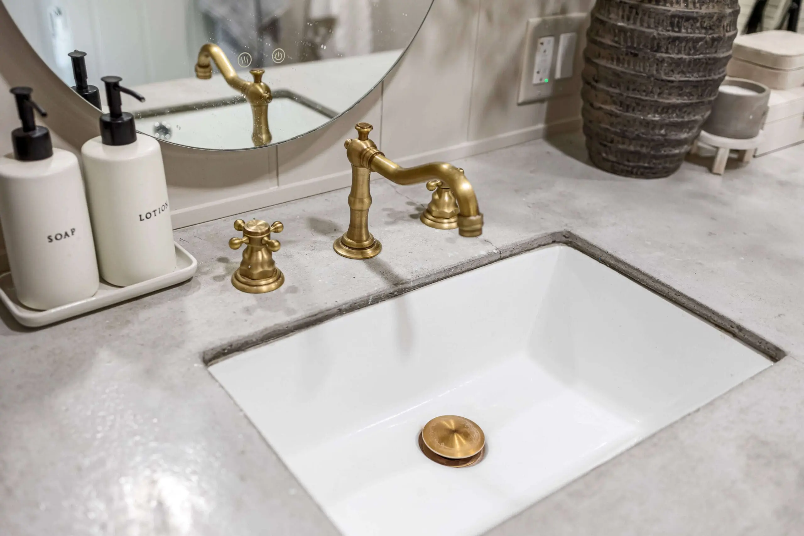 Close-up of a bathroom sink set in a polished concrete countertop, featuring elegant brass fixtures. The countertop is styled with white soap and lotion dispensers in a tray, and a textured vase is visible in the background.