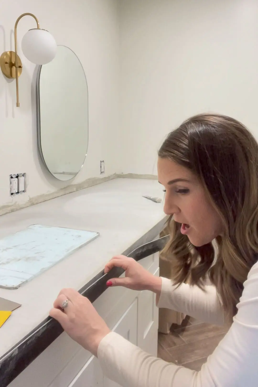 A woman carefully removing the black plastic edge form from the side of a concrete countertop on a bathroom vanity. The finished concrete surface is visible, along with a gold wall sconce and an oval mirror mounted on the wall.