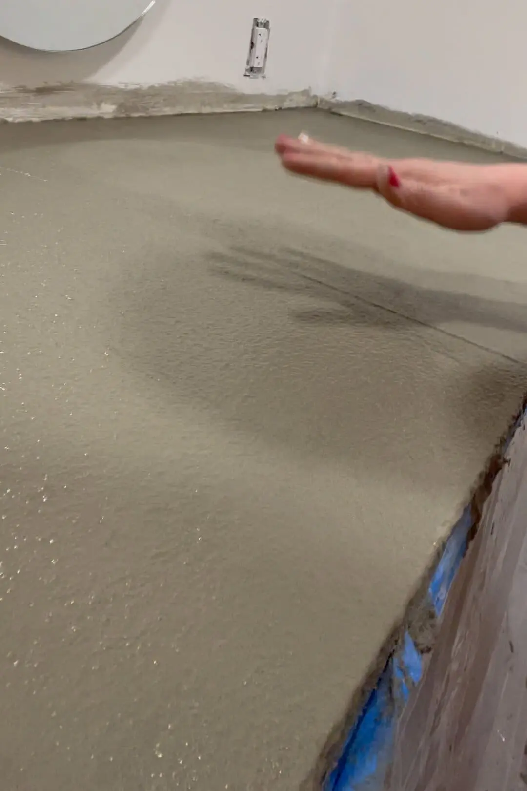 Close-up of a freshly smoothed concrete countertop during the curing process, with a hand lightly gliding over the surface to check for consistency and smoothness. The countertop is part of a bathroom vanity project.