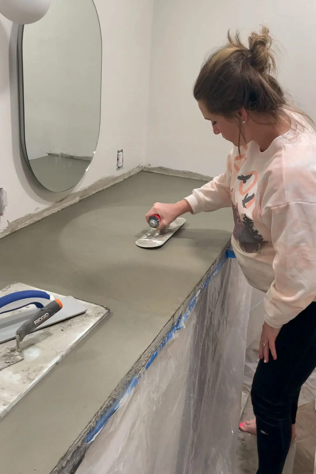 A woman using a finishing trowel to smooth out the surface of a freshly poured concrete countertop on a bathroom vanity. Tools and protective plastic sheeting are visible in the workspace, with an oval mirror mounted on the wall. These DIY countertops are part of a primary bath remodel.