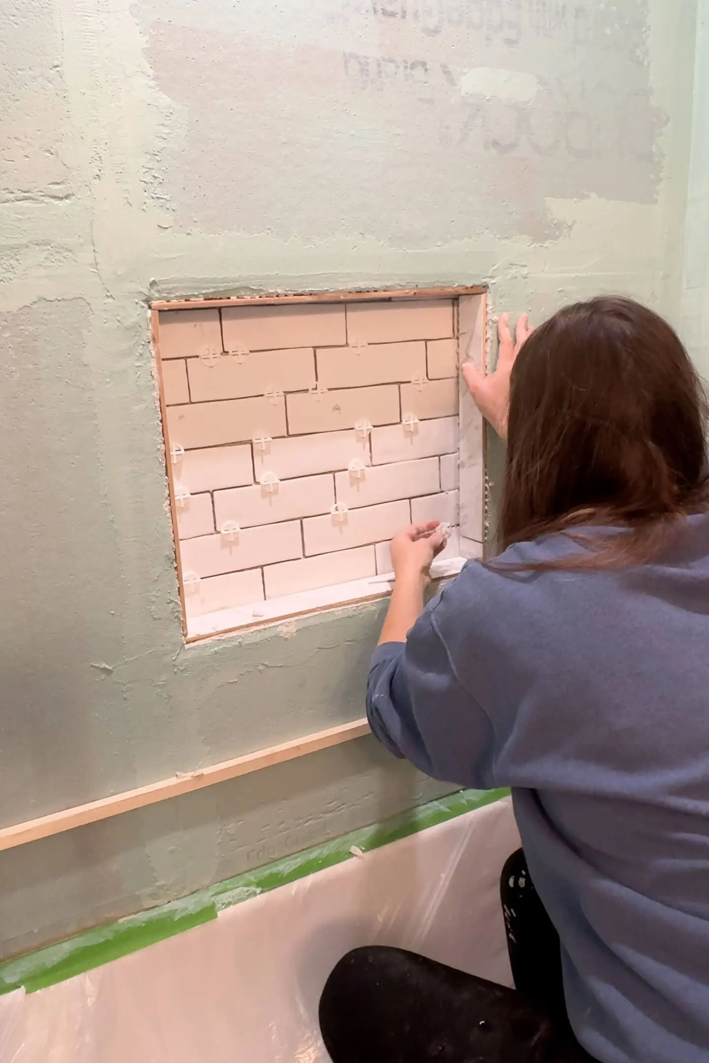 A woman adjusting the placement of white tiles within a recessed niche as part of a tile tub surround. The niche is surrounded by a light green waterproof coating, and tile spacers are used to ensure even spacing.