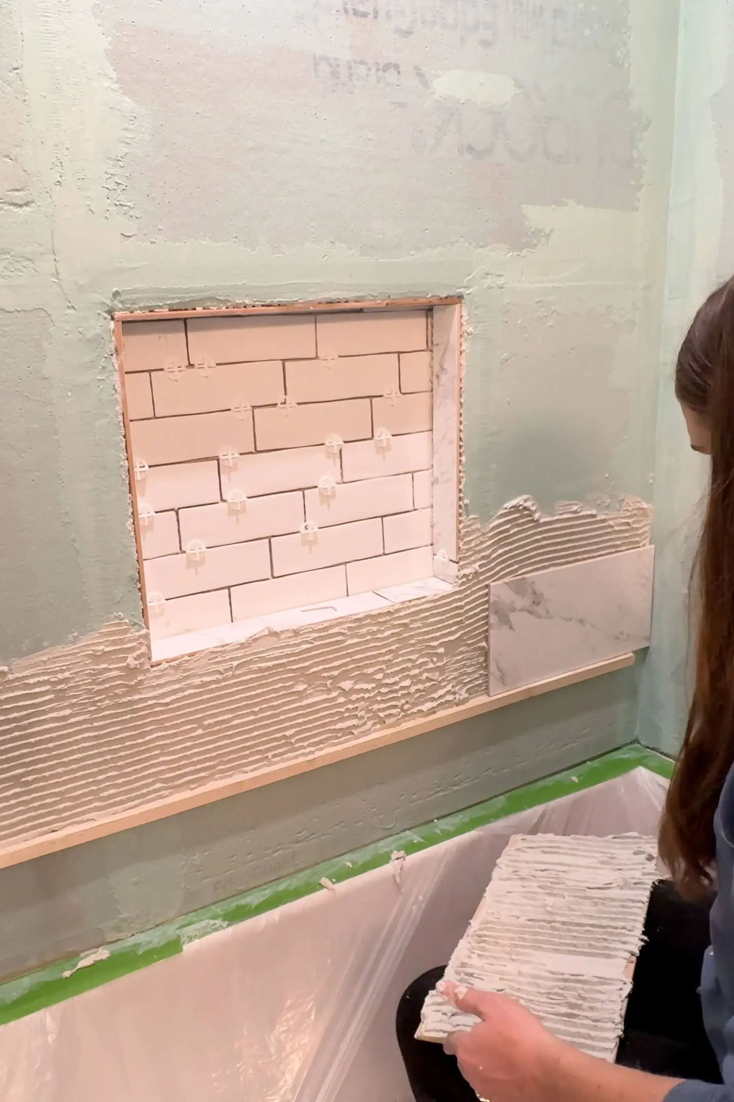 A woman preparing to install marble-style tiles on a tile tub surround. A layer of adhesive mortar is spread across the wall, and the recessed niche is visible, tiled with white tiles in a brick pattern.