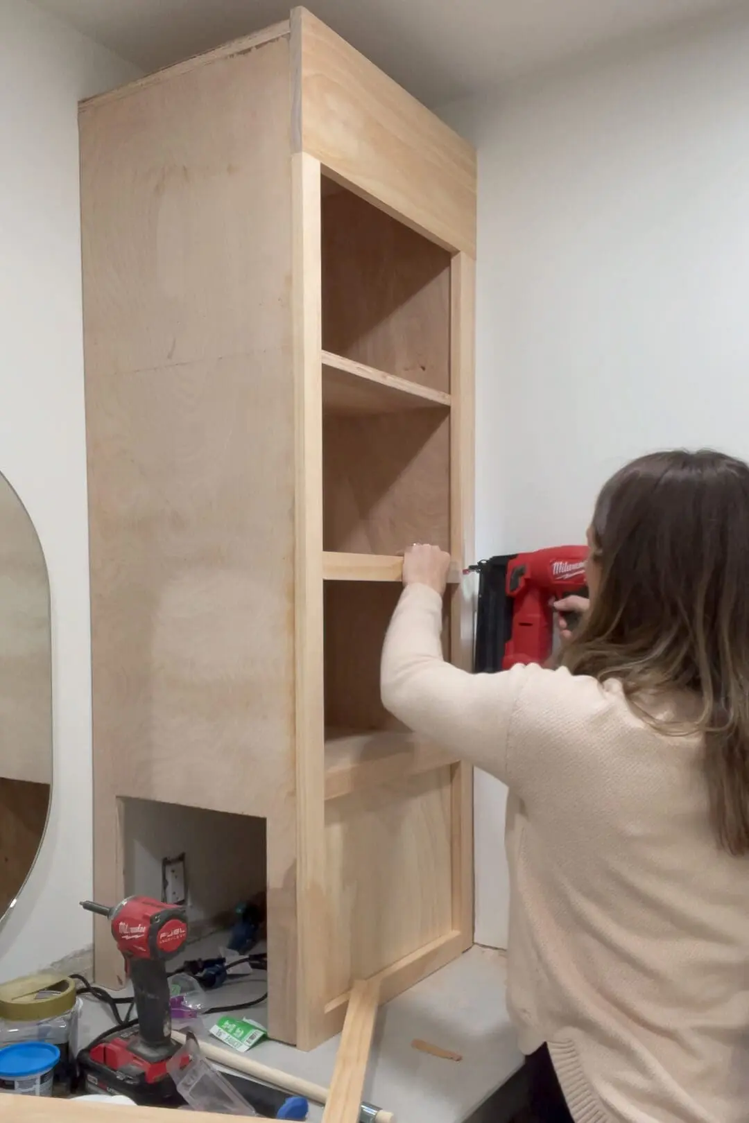 Woman using a red Milwaukee nail gun to secure trim pieces on a custom-built wooden counter cabinet. The cabinet, made from unfinished plywood, is being assembled atop a concrete vanity in a bathroom under construction.