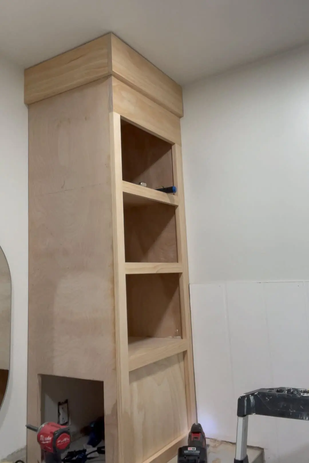 Completed installation of a custom-built wooden counter cabinet with trim detailing. The tall shelving unit is unfinished, with natural wood grain visible, and is built into a bathroom vanity.