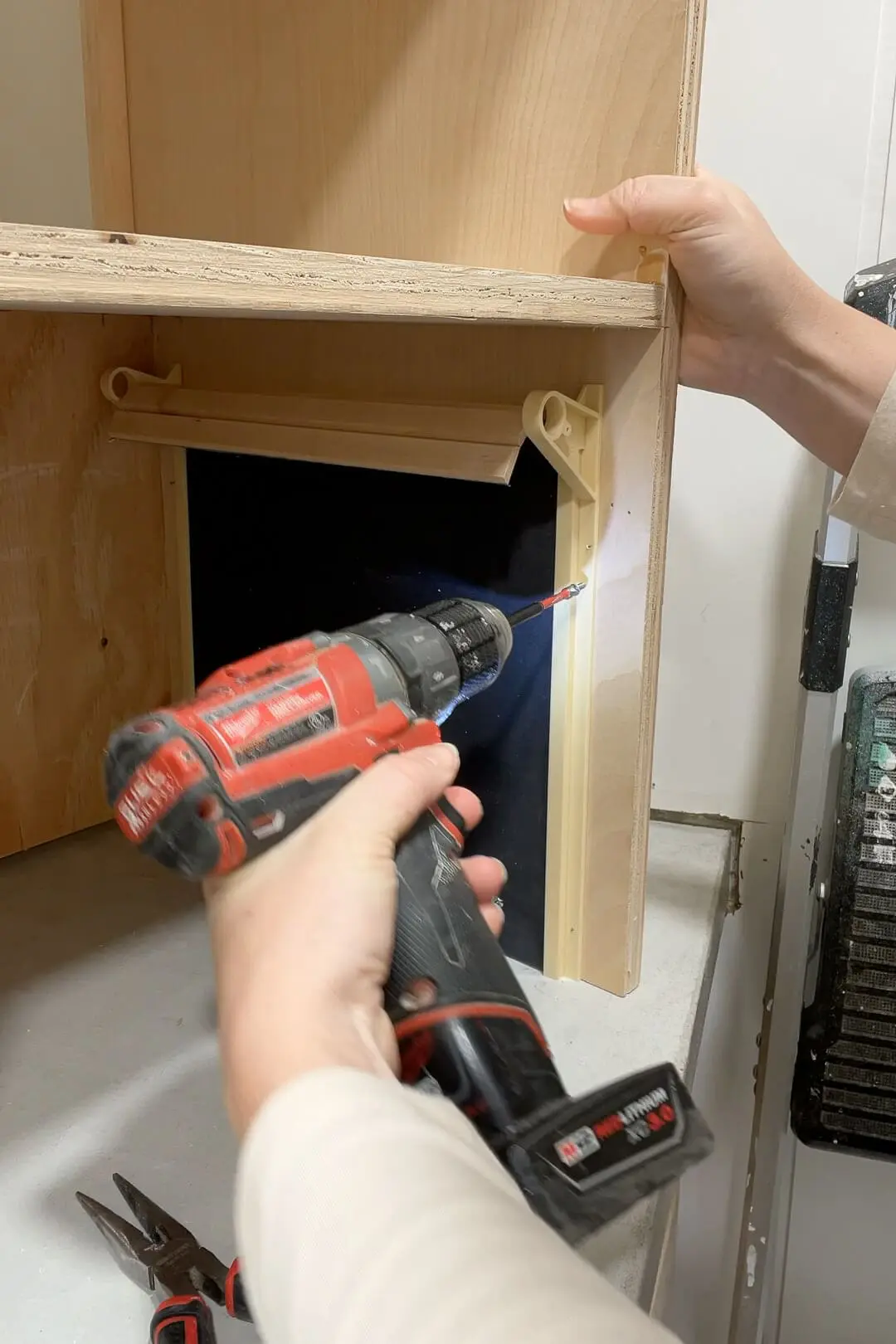 Close-up of hands using a red Milwaukee drill to secure a wooden support piece inside a custom cabinet. The inside features plastic hardware components, indicating a pull-down or roll-up mechanism.