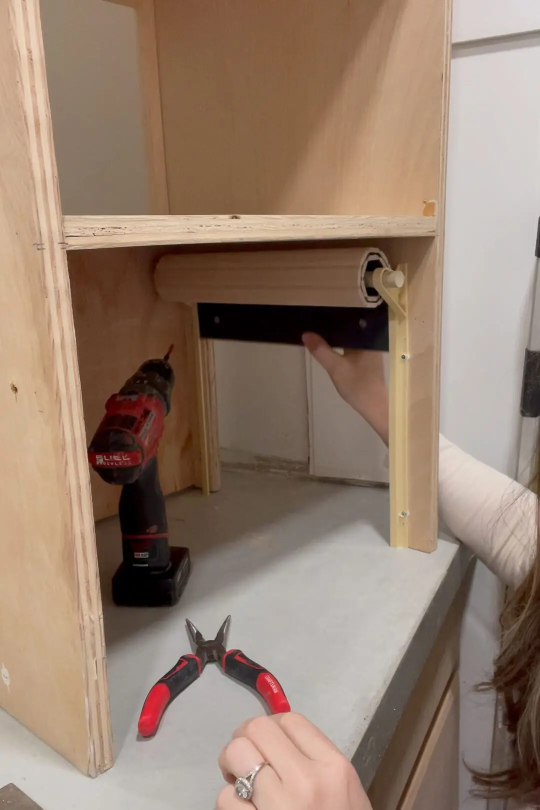 Woman adjusting the tambour door hardware inside a custom-built bathroom cabinet. She tightens screws on the plastic side track that allows the slatted door to slide smoothly.