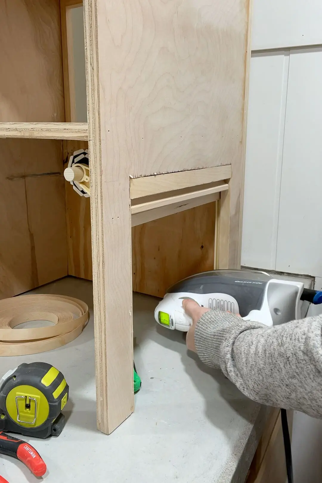 Woman using a household iron to apply edge banding to the unfinished plywood cabinet. A roll of wood veneer edge banding sits nearby, along with a tape measure and pliers on the concrete countertop.