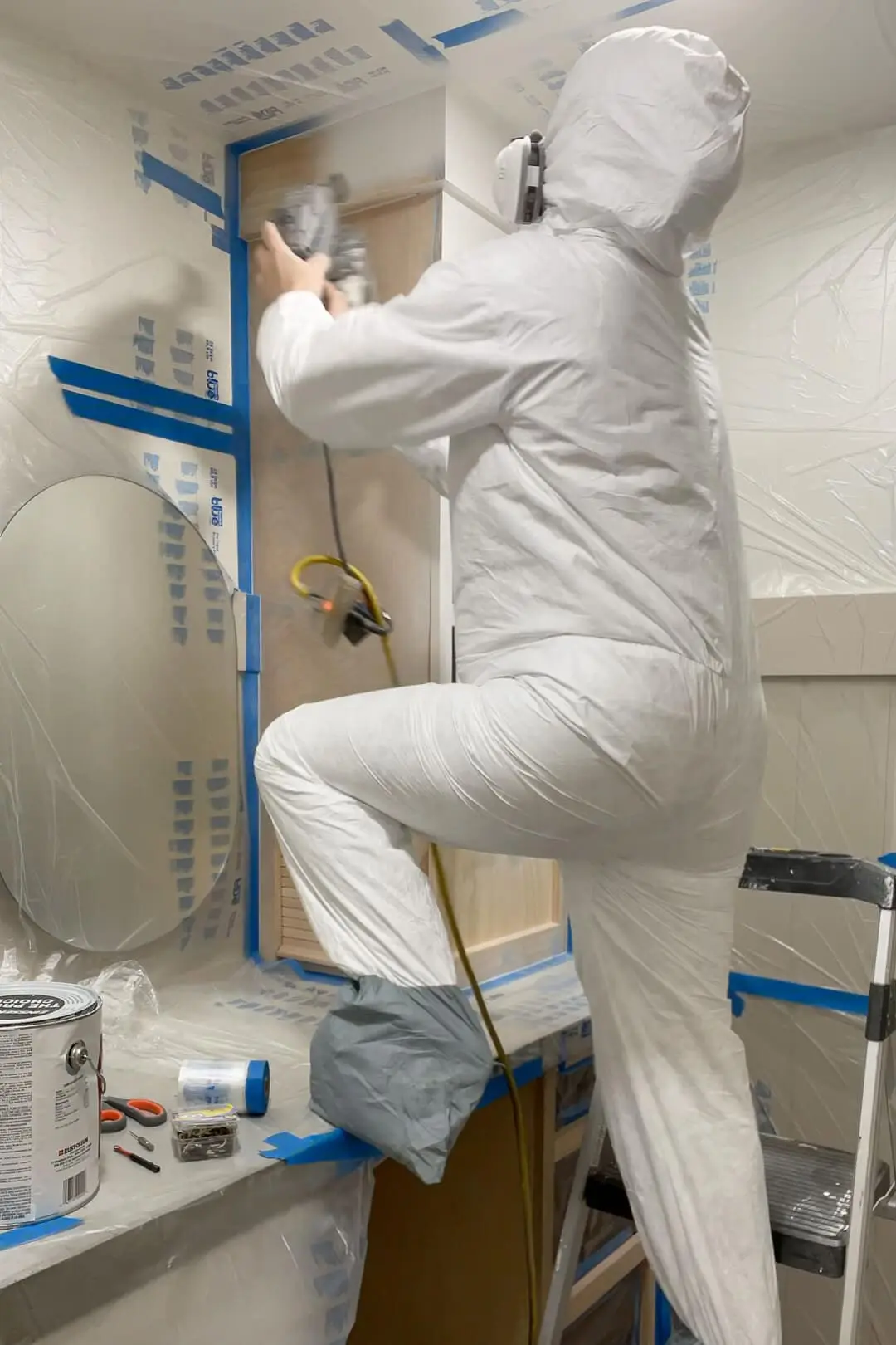 Person in a full-body protective suit and respirator using a paint sprayer to coat the custom-built bathroom cabinet. The workspace is covered in plastic sheeting and painter’s tape to protect surfaces.