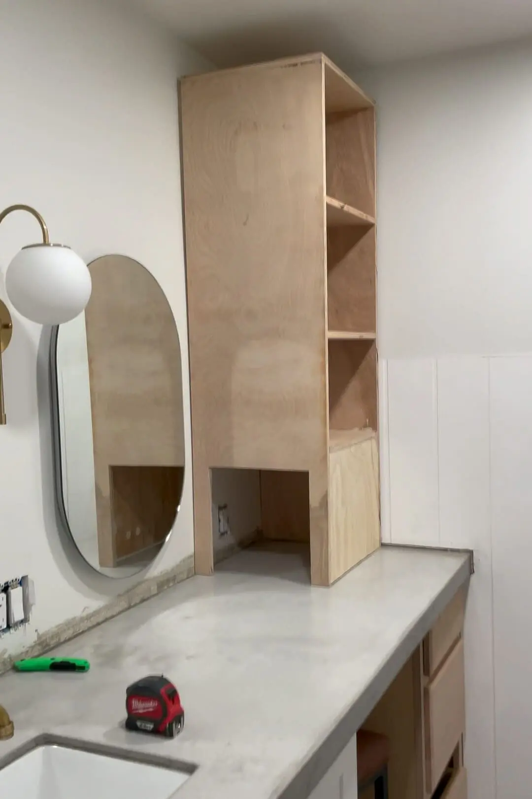 Partially installed custom wooden storage tower sitting on a concrete vanity in a bathroom. An arched mirror, a modern gold sconce, and unfinished cabinetry surround the new addition, highlighting the DIY renovation progress.
