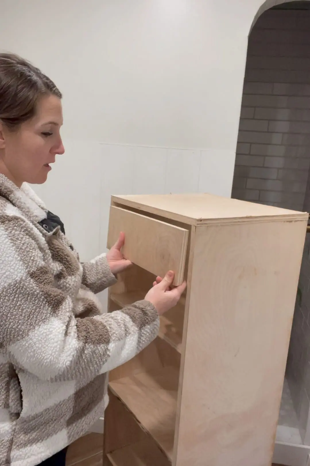 Woman fitting a wooden cabinet door into place on a custom-built storage unit. She holds the door panel up to check the alignment before securing it, wearing a cozy plaid fleece.