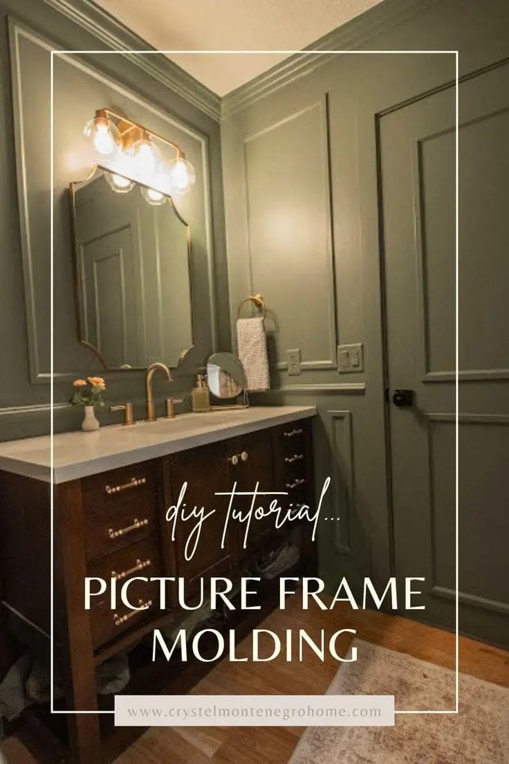 A modern farmhouse bathroom with sage green walls featuring paneled molding, a dark wood vanity with brass hardware, a white countertop, and gold fixtures. The room is styled with a vintage gold-framed landscape painting, wooden wall hooks, and a small floating wooden shelf holding decorative jars and greenery.