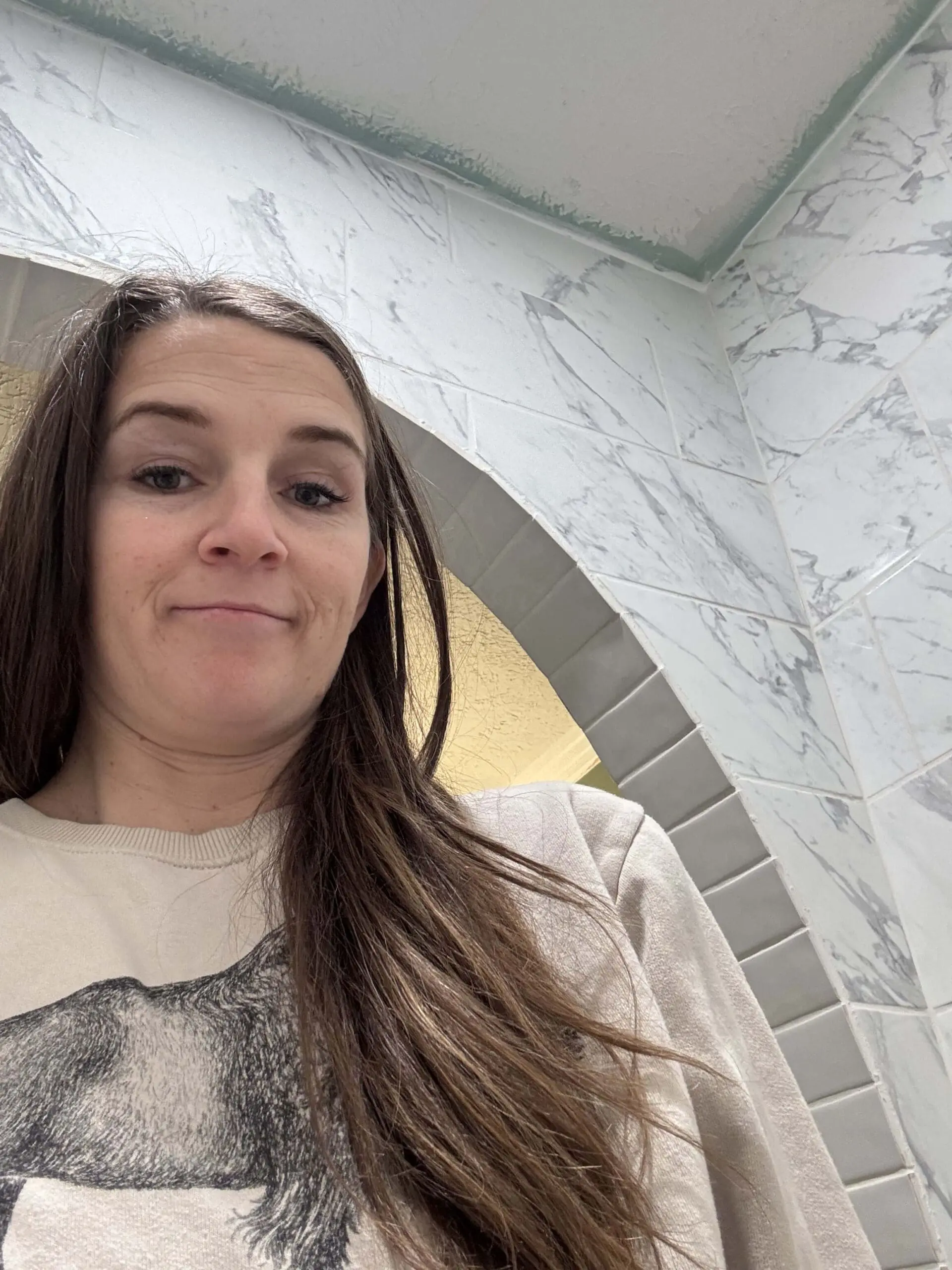 Woman in a casual cream-colored shirt with long brown hair, taking a selfie in a bathroom featuring a tiled marble-look wall with an arched design and a painted ceiling edge.