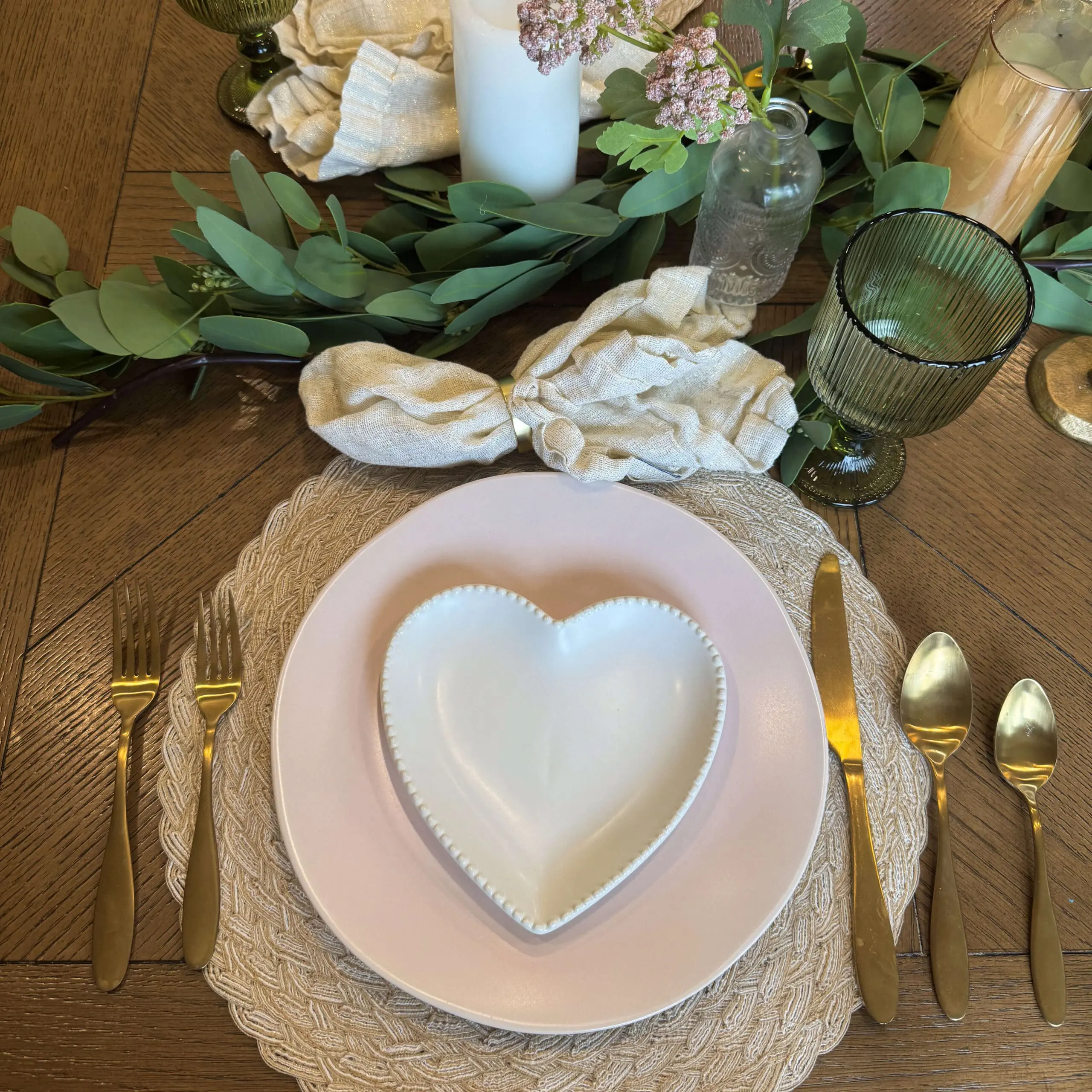 A close-up of a romantic Valentine's Day 2025 table setting featuring a white heart-shaped plate on top of a soft pink dinner plate, placed on a textured woven placemat. The table is decorated with gold flatware, linen napkins tied with golden rings, eucalyptus greenery, and a green glass goblet.