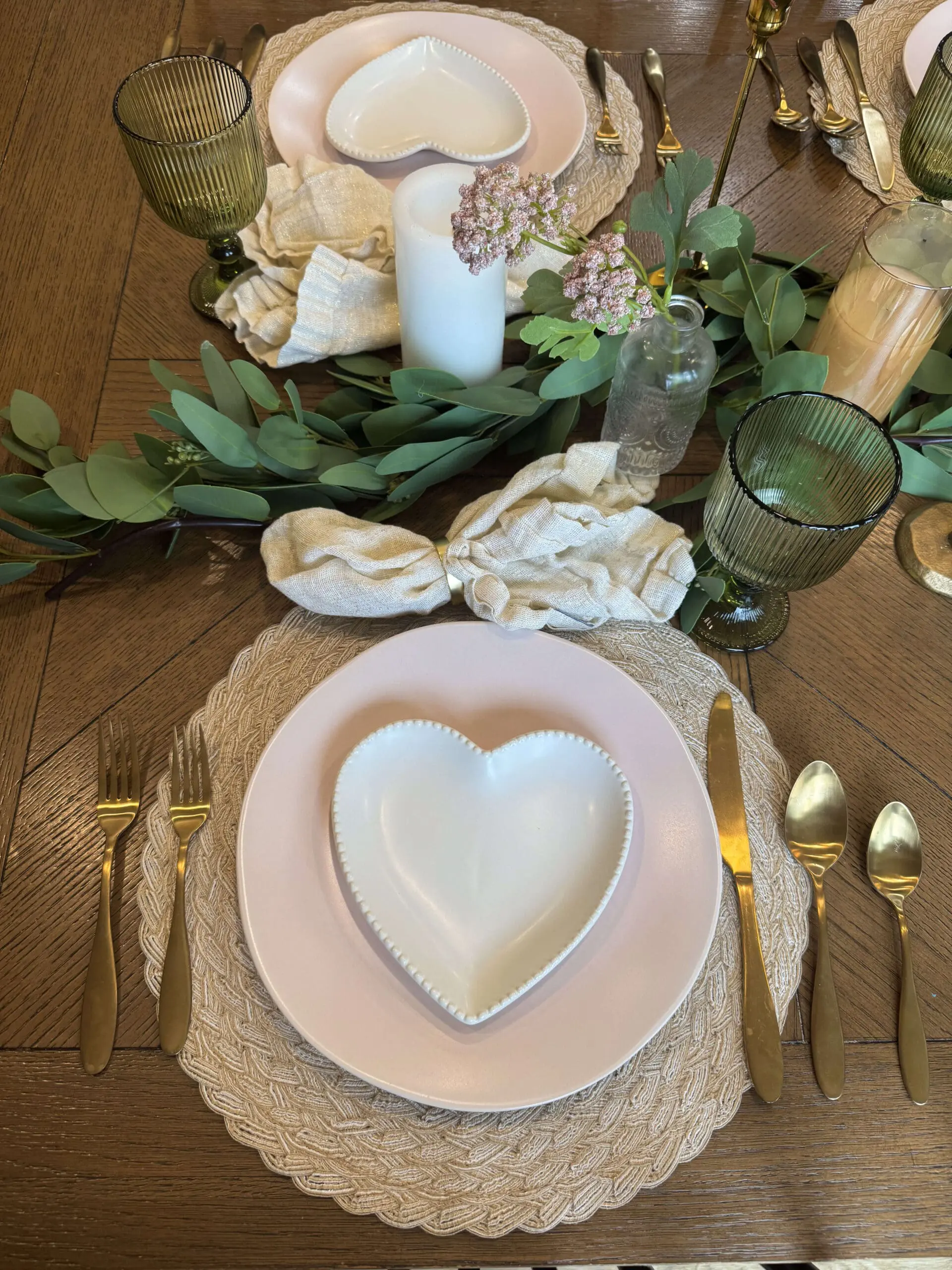 A close-up of a romantic Valentine's Day 2025 table setting featuring a white heart-shaped plate on top of a soft pink dinner plate, placed on a textured woven placemat. The table is decorated with gold flatware, linen napkins tied with golden rings, eucalyptus greenery, and a green glass goblet.