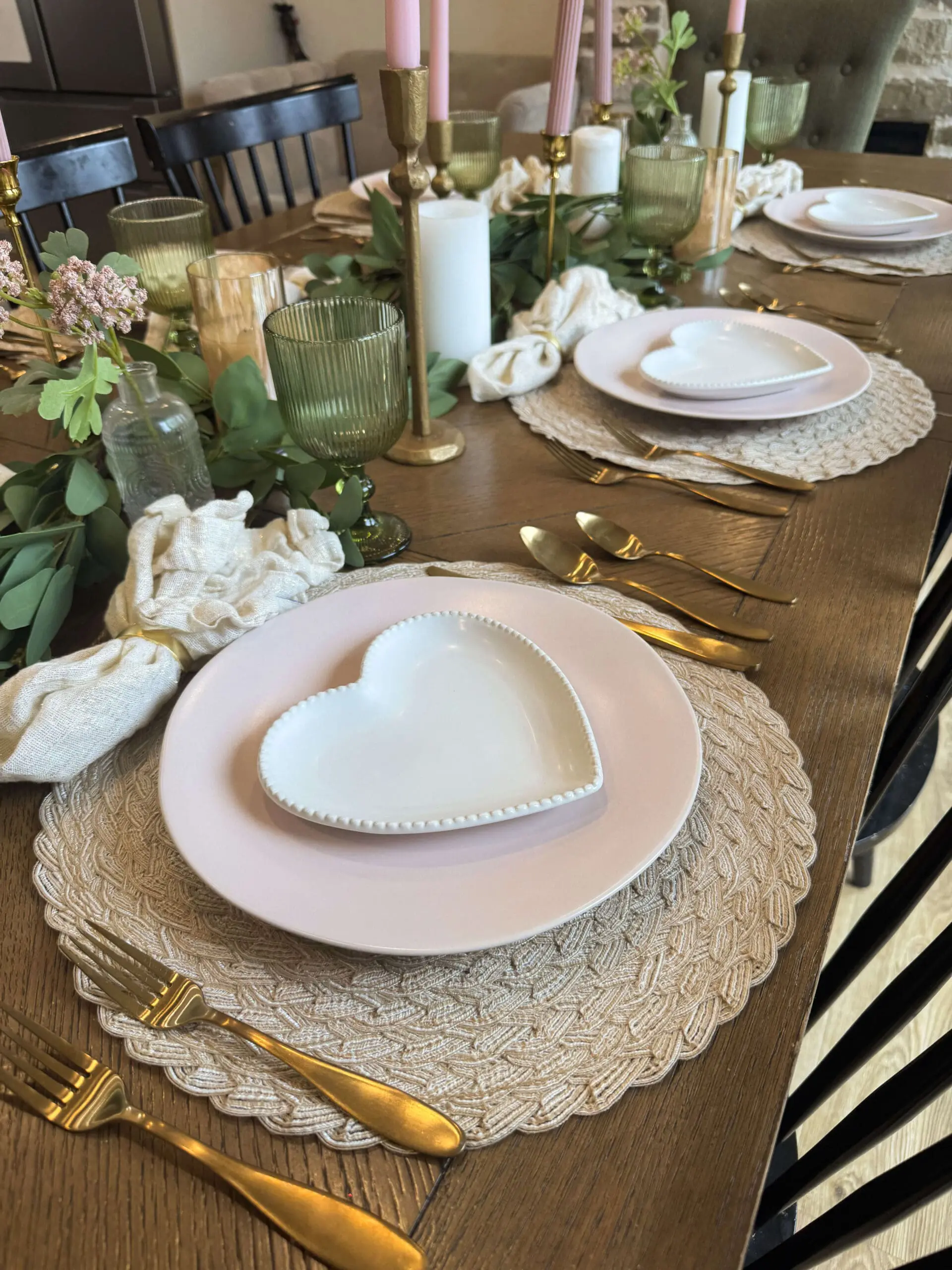 An angled view of my Valentine's Day 2025 dining table showcasing delicate heart-shaped plates on pink dishes, gold flatware, woven placemats, and green ribbed goblets. The table is adorned with eucalyptus greenery, blush pink candles, and soft linen napkins for a charming and inviting setting.