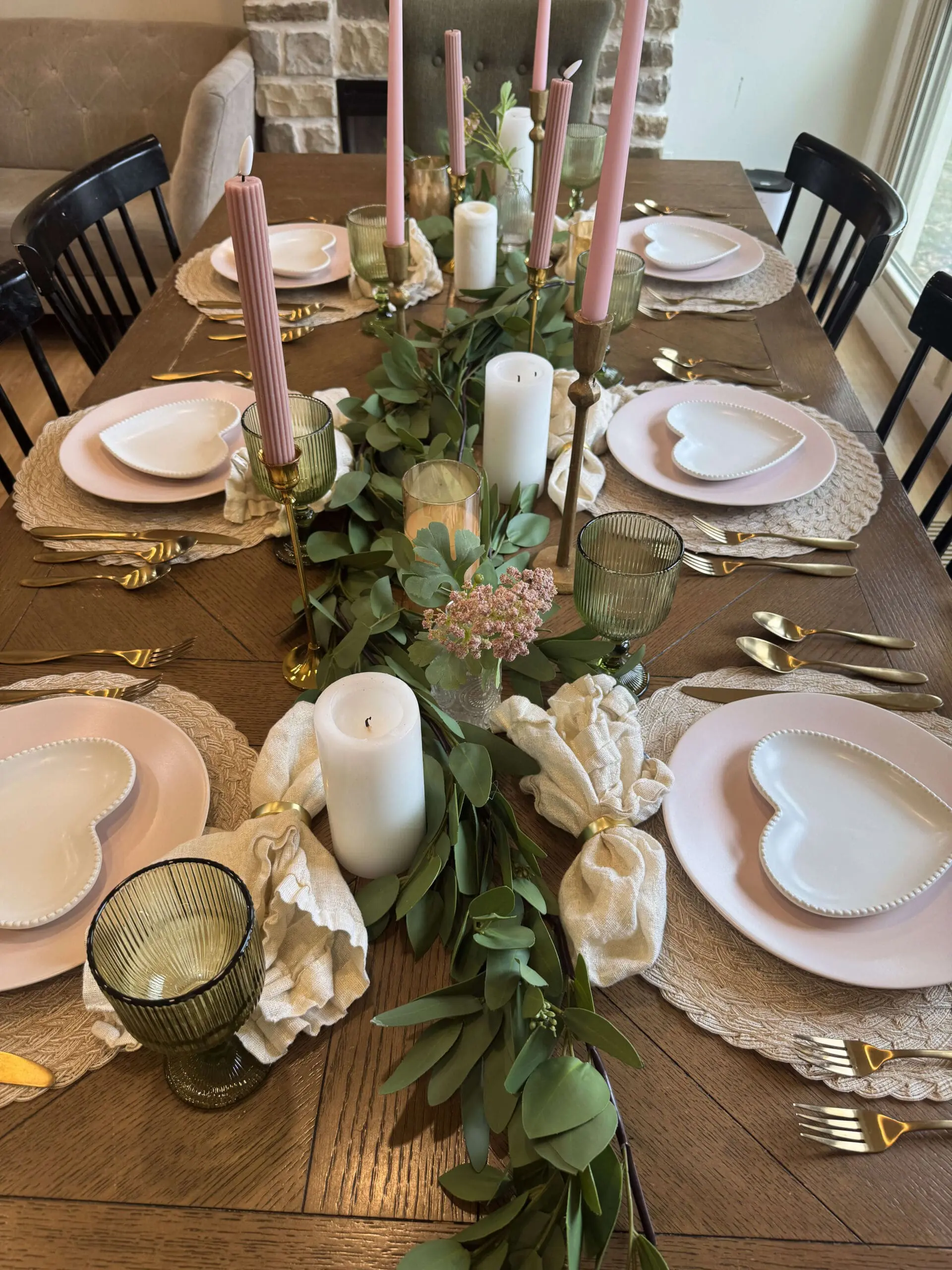 A beautifully styled Valentine's Day 2025 table set for a romantic gathering, featuring white heart-shaped plates layered on pink dinner plates. The centerpiece includes lush eucalyptus garlands, tall pink tapered candles in gold holders, and white pillar candles, creating an elegant and cozy atmosphere.