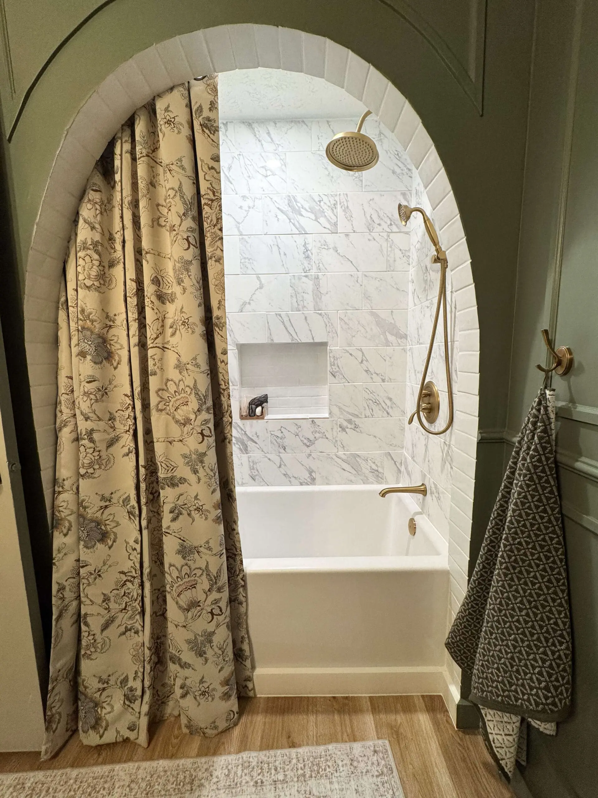A bright and elegant bathtub alcove with white marble tiles, brass shower fixtures, and a built-in niche for bath essentials. The space is framed by a green arched wall and complemented by a floral curtain and patterned bath mat.