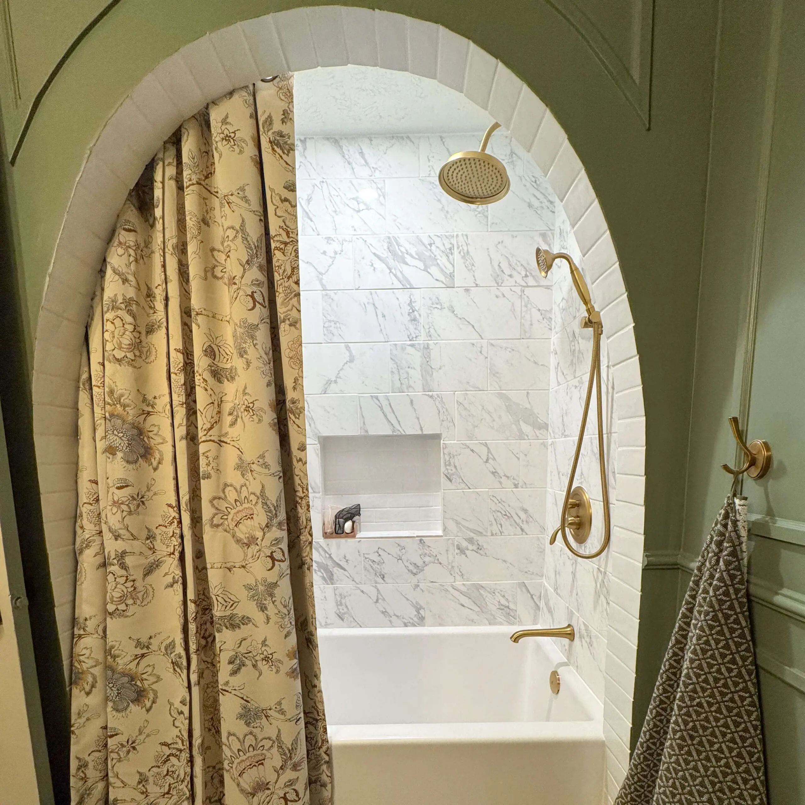 A bathtub area framed by a sage green arched bathtub alcove with white subway tiles. The shower features brass fixtures, including a rainfall showerhead and handheld sprayer, set against white marble tiles with gray veining. A floral curtain and textured towel add a cozy touch.