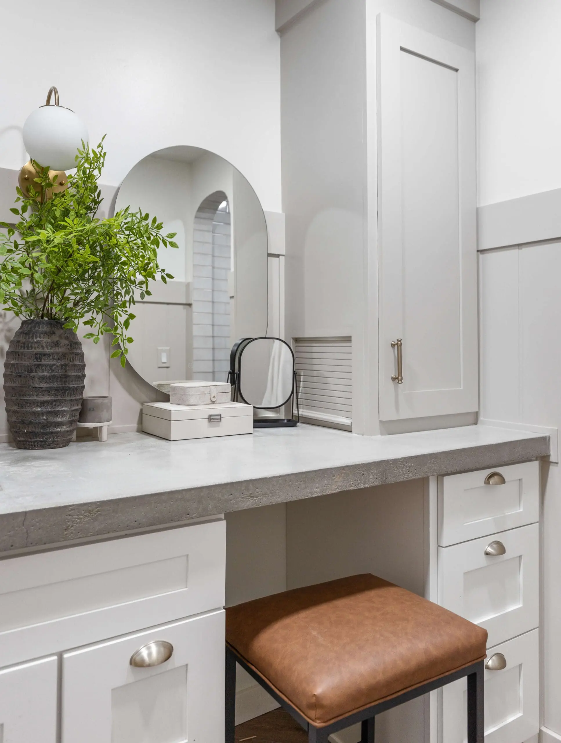 Well-organized bathroom vanity featuring a concrete countertop, white cabinets with brushed gold handles, and a built-in storage tower. A brown leather stool with a black metal frame is tucked neatly under the counter, offering a stylish seating option for this serene and functional space.