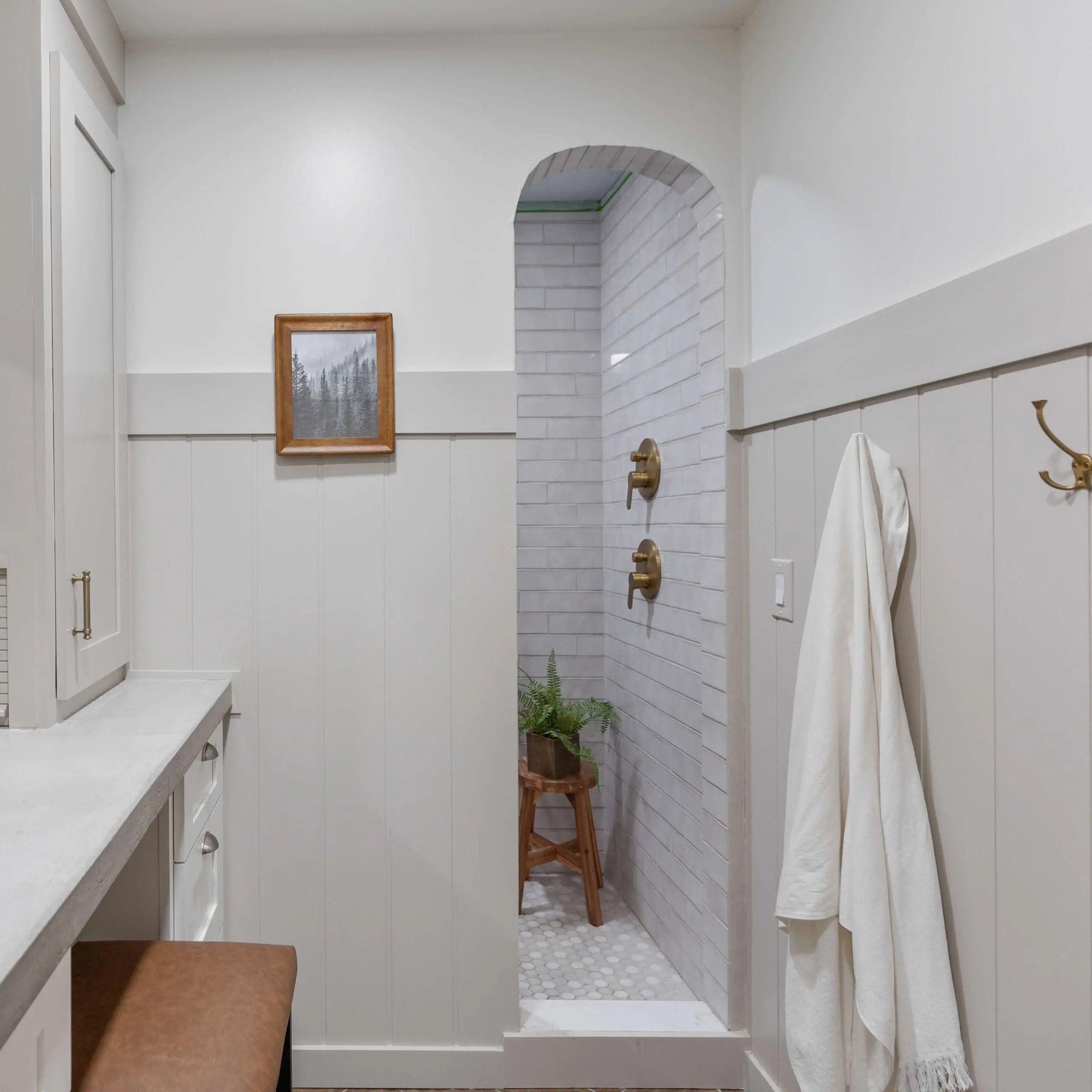 A beautifully designed neutral-toned bathroom featuring an arched walk-in shower with soft gray subway tiles, brass shower fixtures, and a cozy wooden stool with a potted green fern. The walls are adorned with white board and batten wainscoting, a simple black-and-white framed landscape print, and a plush white towel hanging on a brass hook.