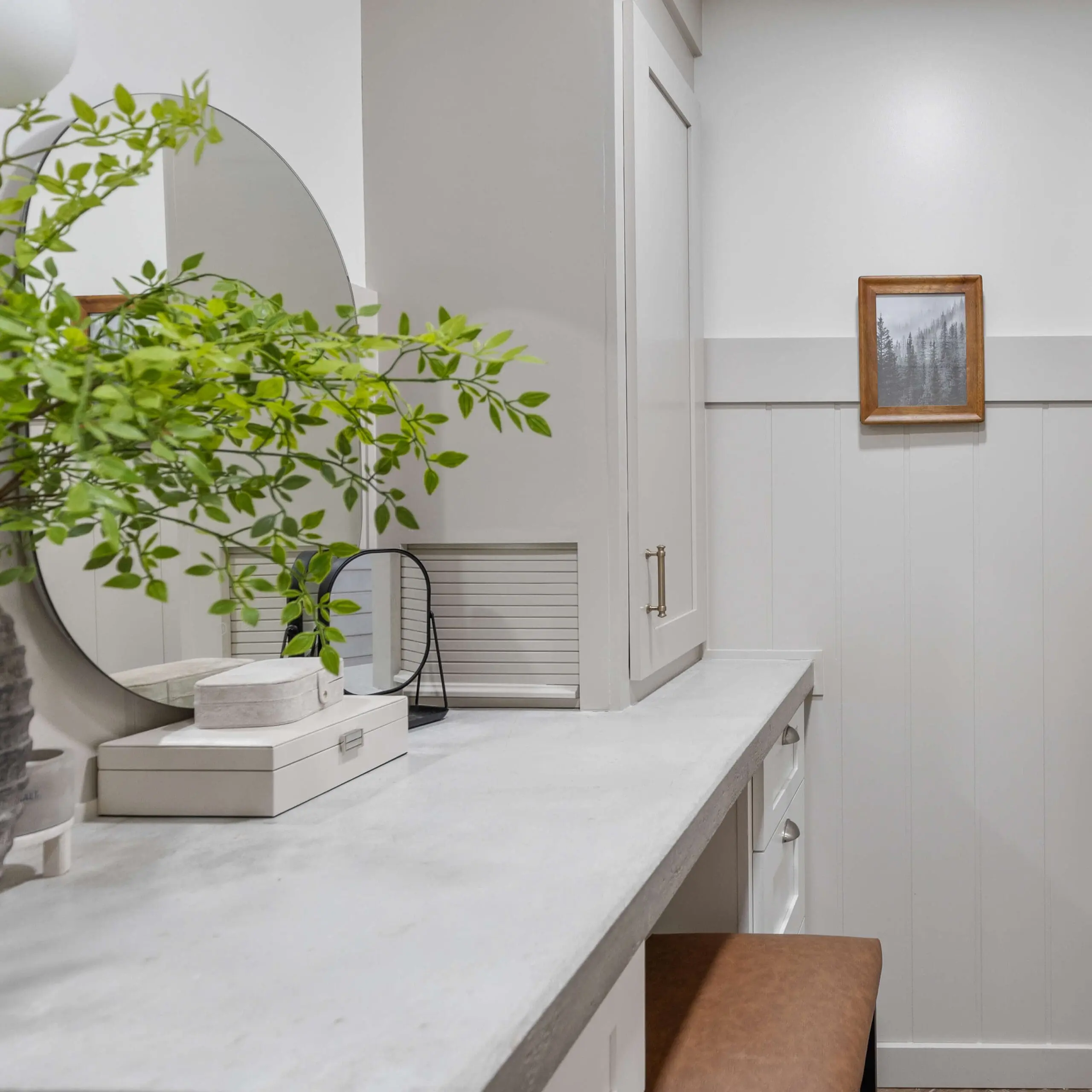 A functional bathroom counter area with concrete countertops and a soft leather stool tucked underneath. Features include greenery in a decorative vase, small storage boxes, and a round mirror reflecting the serene decor.