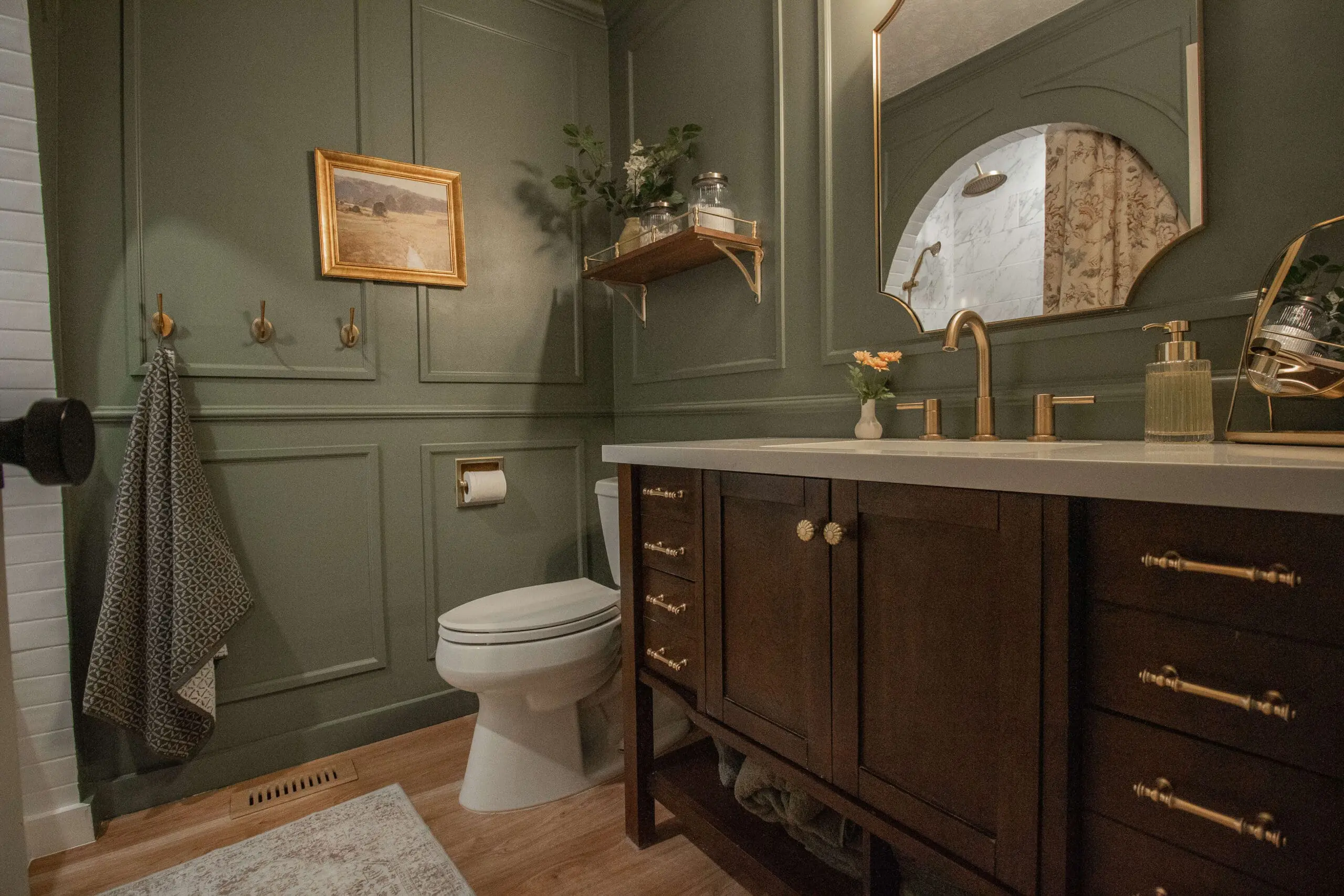 A modern farmhouse bathroom with sage green walls featuring paneled molding, a dark wood vanity with brass hardware, a white countertop, and gold fixtures. The room is styled with a vintage gold-framed landscape painting, wooden wall hooks, and a small floating wooden shelf holding decorative jars and greenery.